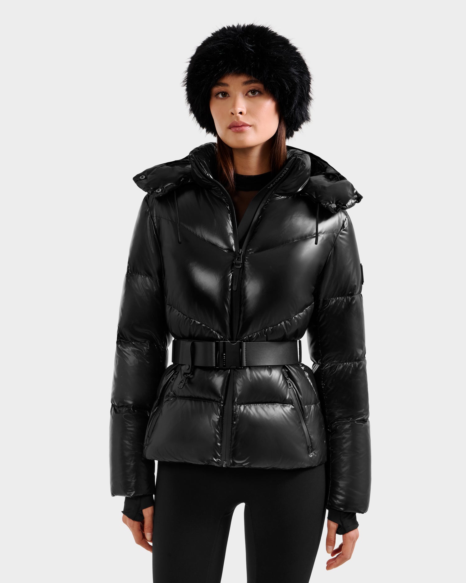 A woman in the Rudsak CHRISTELLE S - L125544 Black shiny winter jacket with a belt, black leggings, and a black furry hat stands against a plain light gray background.