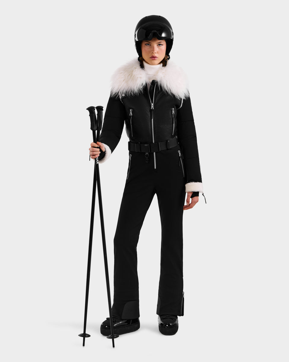 A woman wears the CHYNTIA - 8125611 Black ski suit by Rudsak, with a white fur collar, black gloves, boots, and helmet. She stands facing forward against a white background. Pre-order now; ships by November 30th.