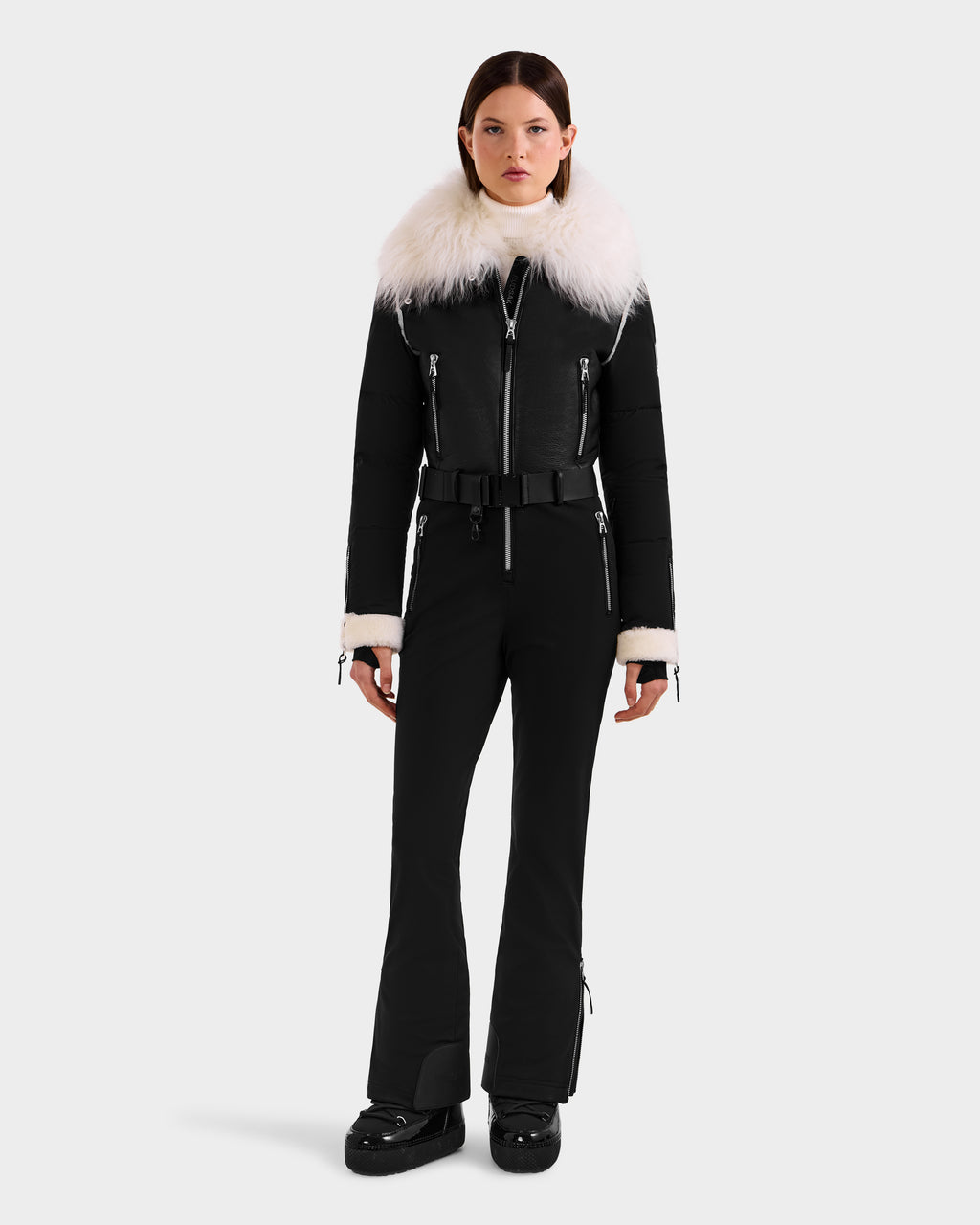 A woman faces forward in the Rudsak CHYNTIA - 8125611 Black fitted winter jumpsuit with a wide white faux fur collar and cuffs, black belt, and boots. Now available for pre-order; ships by November 30th.