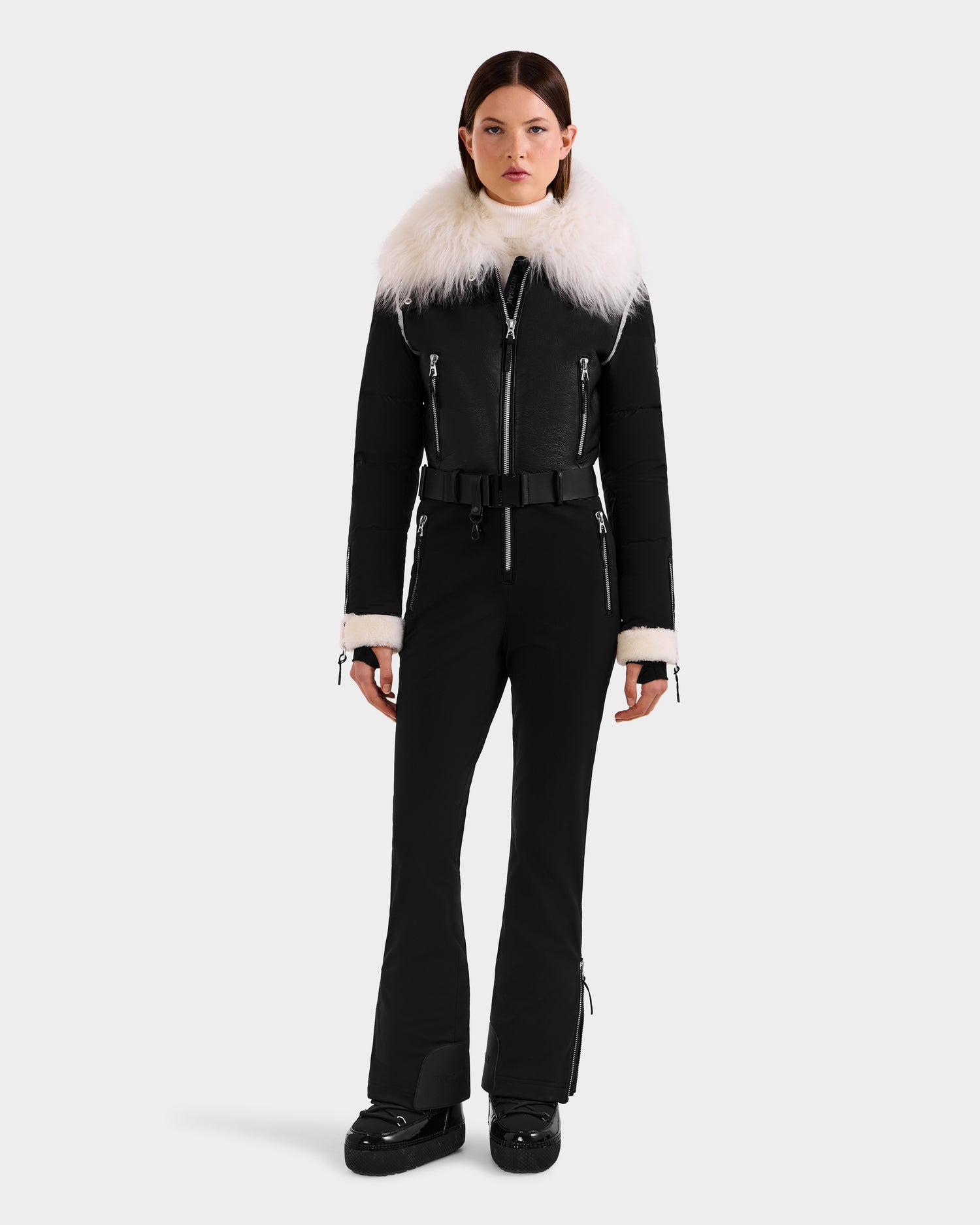 A woman faces forward in the Rudsak CHYNTIA - 8125611 Black fitted winter jumpsuit with a wide white faux fur collar and cuffs, black belt, and boots. Now available for pre-order; ships by November 30th.