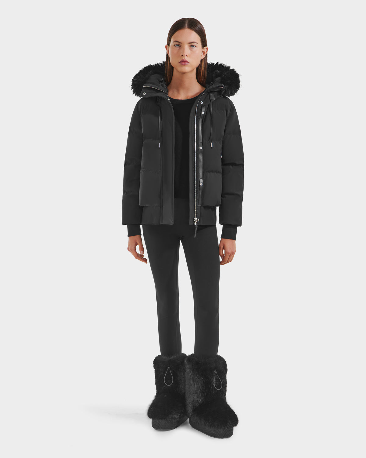 A woman stands facing forward in the Rudsak CIEYNA - 8125579 Black down-filled bomber with faux fur hood trim, paired with black leggings and black furry boots against a plain white background.