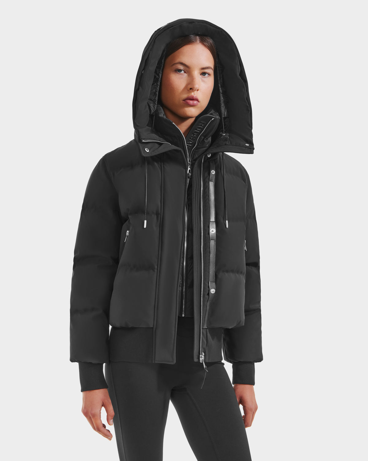 A woman wears the Rudsak CIEYNA - 8125579 Black down-filled bomber with recycled weather-resistant shell and zip details, paired with black pants, standing against a plain white background.