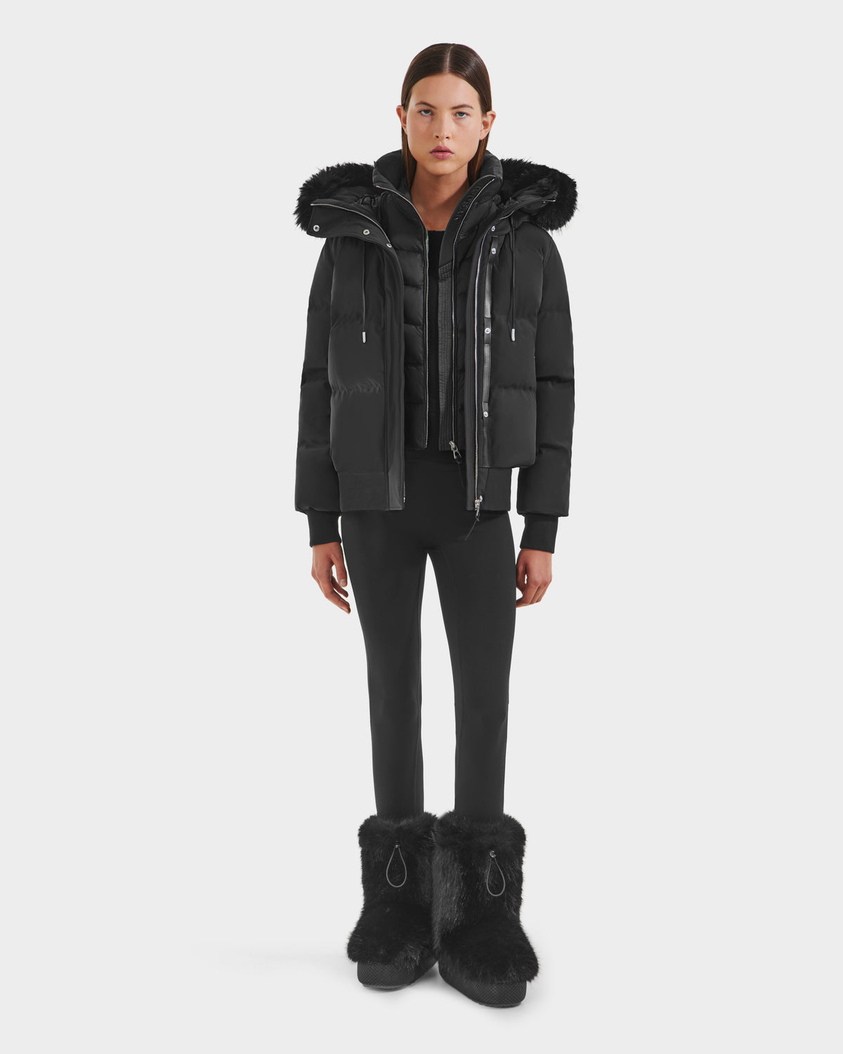 A person stands facing forward, wearing the Rudsak CIEYNA - 8125579 Black down-filled bomber jacket with a faux fur hood trim, black pants, and large black fur boots against a plain white background.