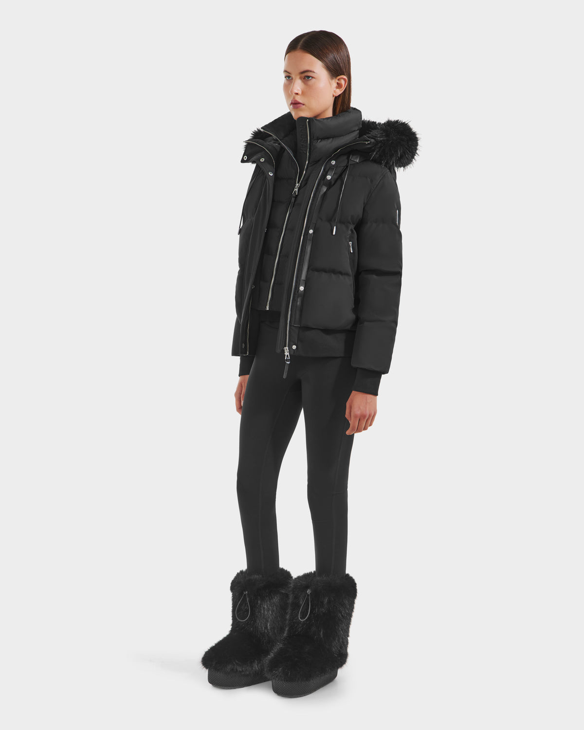 A woman faces forward against a white background, wearing the Rudsak CIEYNA - 8125579 Black puffer jacket with a recycled weather-resistant shell and faux fur hood trim, paired with black leggings and large black furry boots.