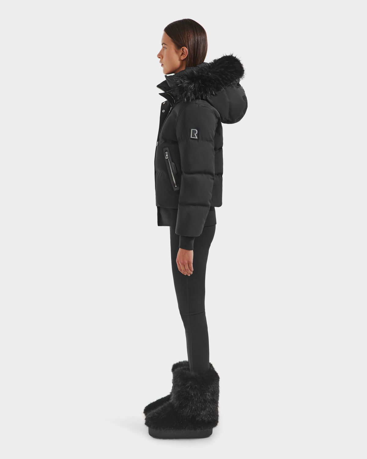 A woman stands in profile against a white background, wearing the Rudsak CIEYNA - 8125579 Black down-filled bomber with faux fur hood trim, paired with black leggings and furry black boots.