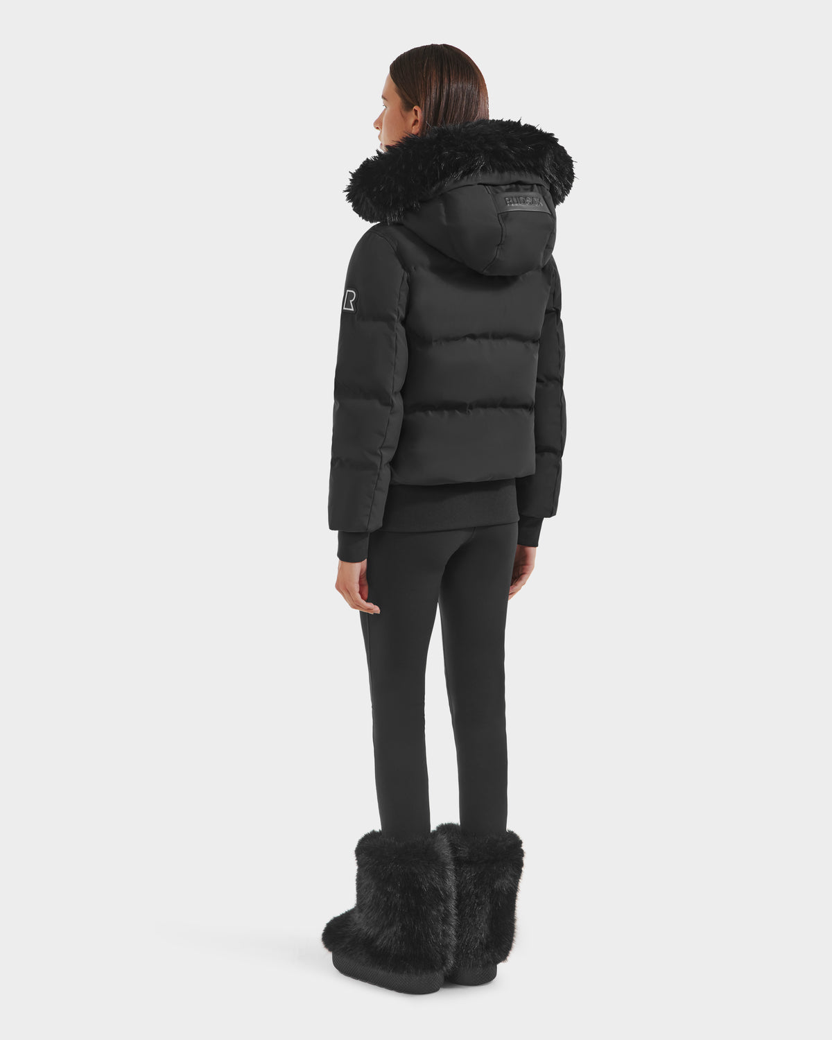 A person stands facing away, wearing the Rudsak CIEYNA - 8125579 Black down bomber with faux fur hood trim, paired with black leggings and tall black furry boots against a plain light background.