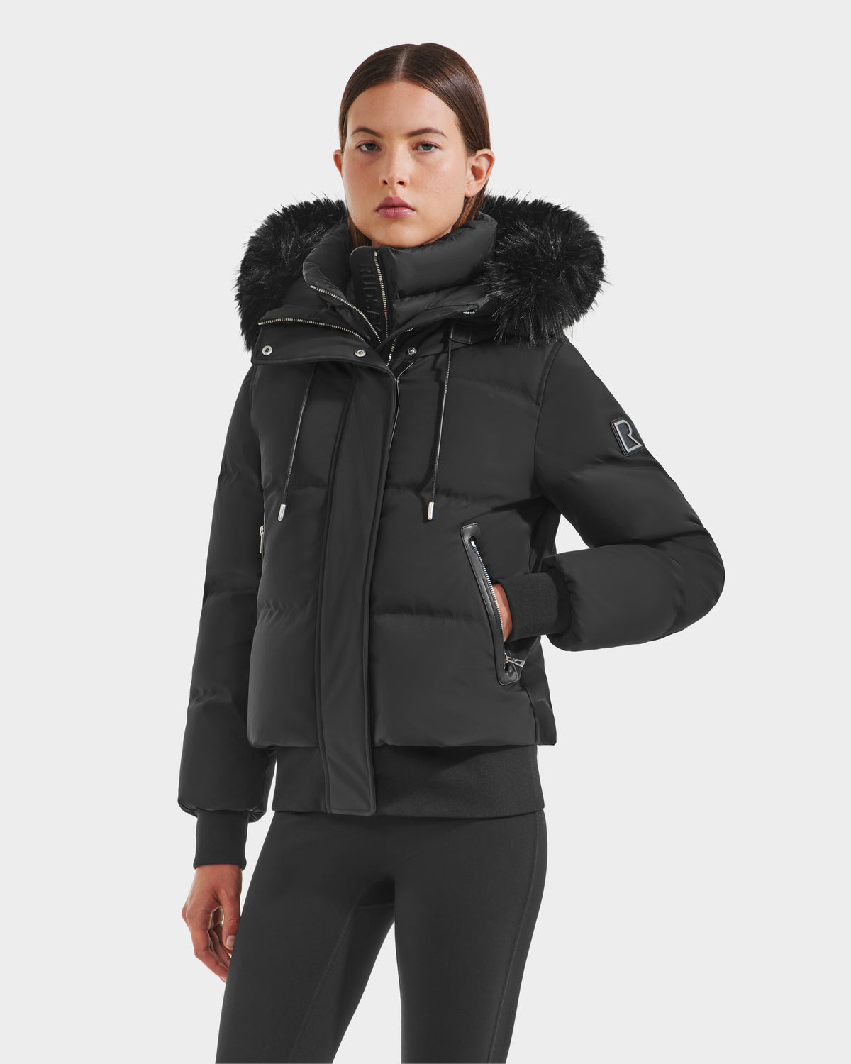 A woman wears the Rudsak CIEYNA - 8125579 Black down-filled bomber with a faux fur hood trim and high collar, paired with black pants, standing against a plain light background.