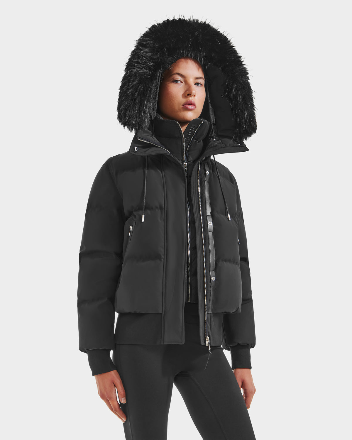 A person wearing the Rudsak CIEYNA - 8125579 Black down-filled bomber with a large faux fur hood trim, black pants, and a zippered inner layer stands against a plain white background.
