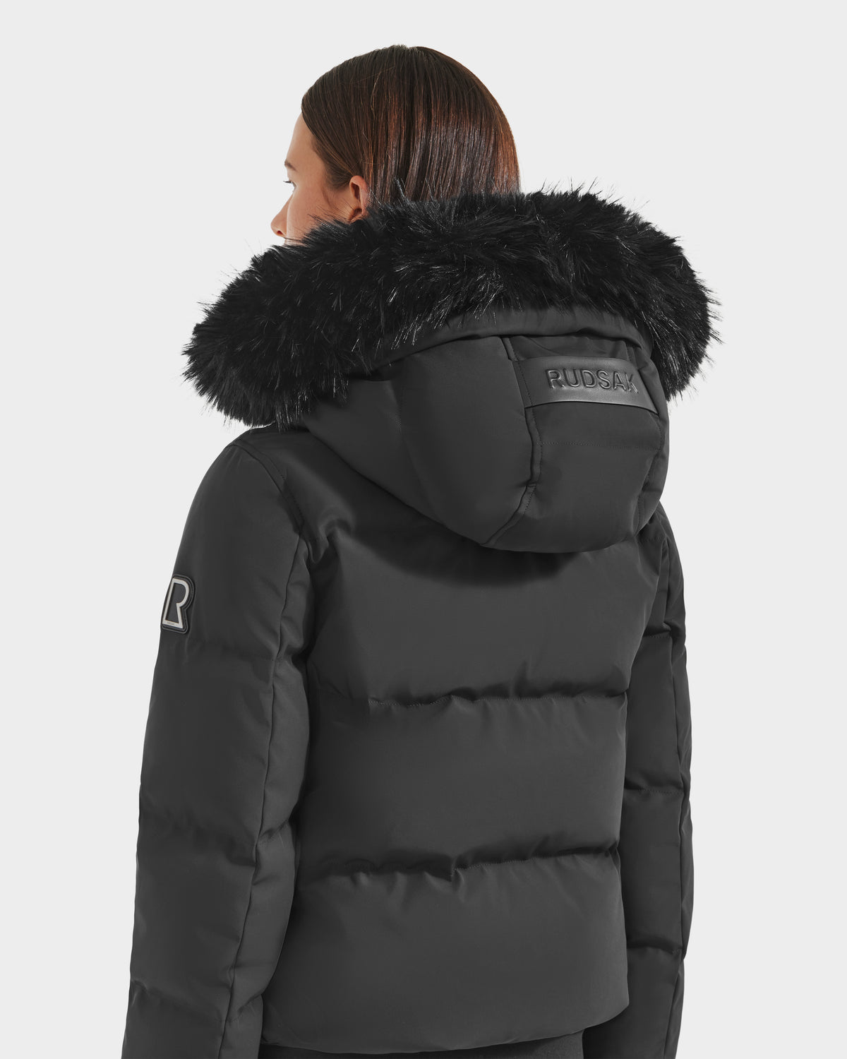 A person with straight brown hair wears the Rudsak CIEYNA - 8125579 Black down-filled bomber, featuring a large faux fur trim, logo patches on the hood and sleeve, and a recycled weather-resistant shell, facing away from the camera.