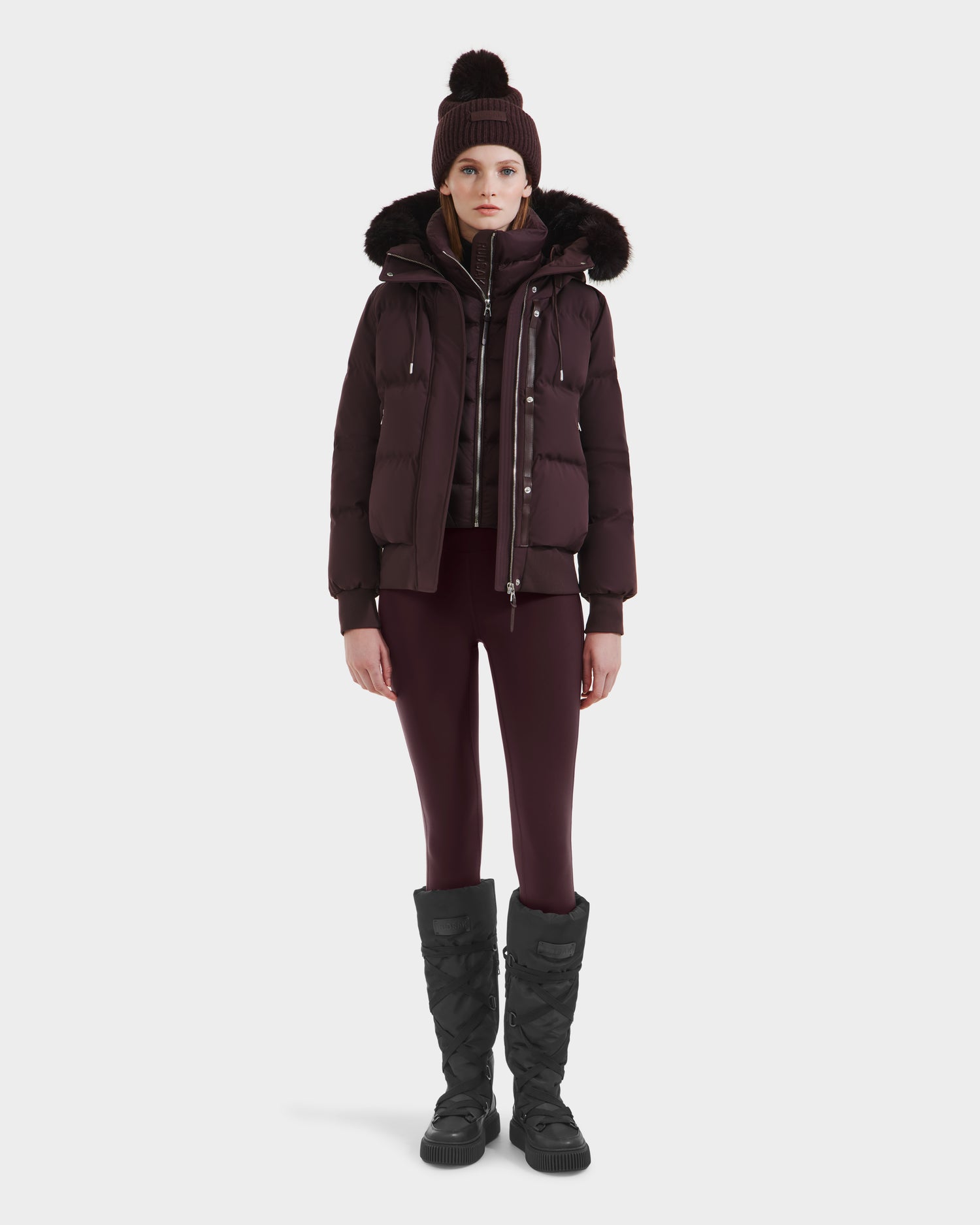 A person wears the Rudsak CIEYNA - 8125579 Bordeaux weather-resistant winter jacket with fur-trimmed hood, paired with a matching hat, plaid sweater, leggings, and black quilted boots, standing against a plain white background.