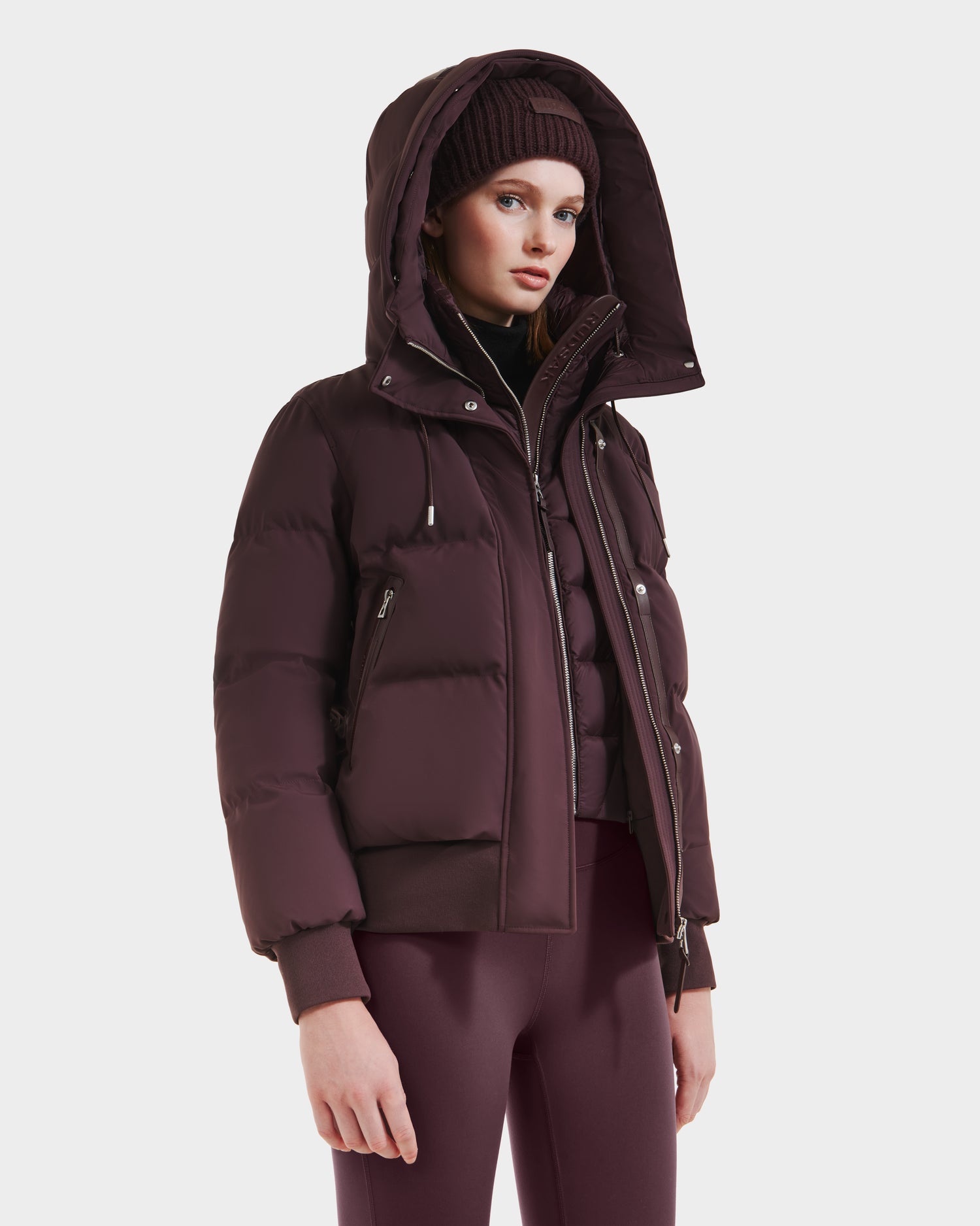 A woman in the Rudsak CIEYNA - 8125579 Bordeaux puffer jacket with hood, paired with a matching knit beanie and fitted leggings, stands against a plain white background.