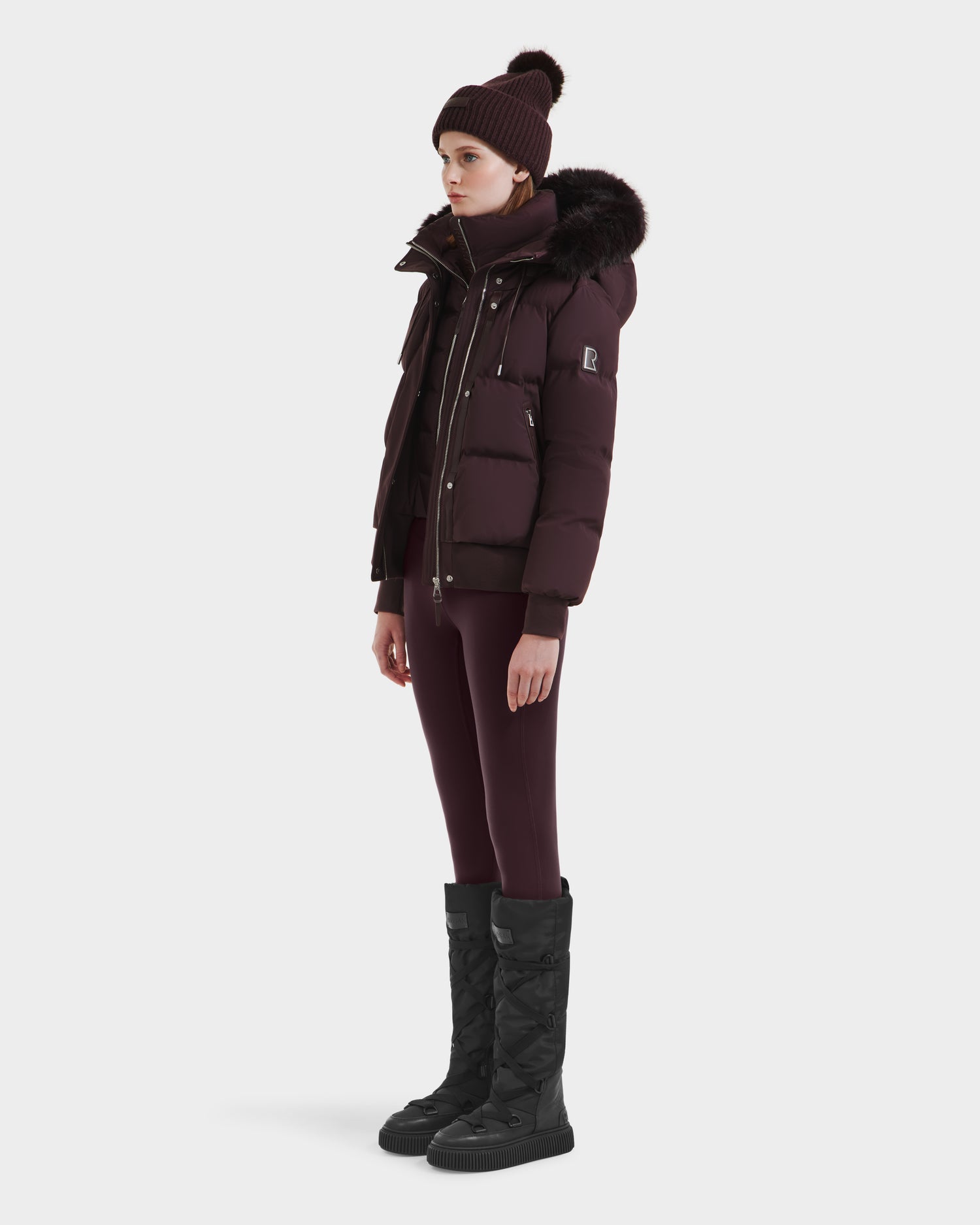 A person stands sideways in the Rudsak CIEYNA - 8125579 Bordeaux weather-resistant puffer jacket with a fur-trimmed hood, styled with matching leggings, a knit beanie, and black quilted boots against a white background.