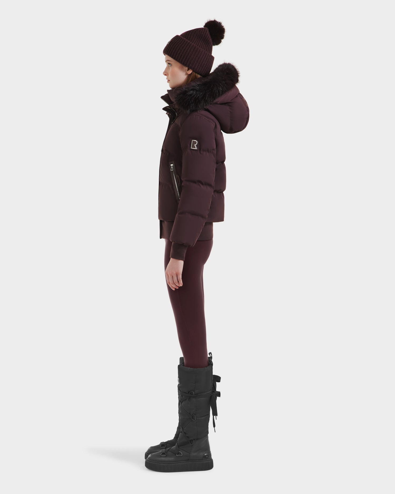 A person in profile wears the Rudsak CIEYNA - 8125579 Bordeaux weather-resistant puffer with a fur-trimmed hood, matching leggings, a pom-pom knit hat, and black boots against a white background.