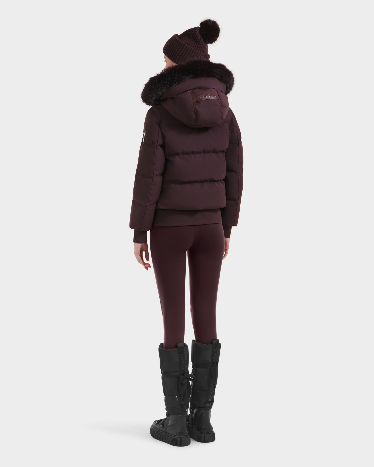 A person faces away in the Rudsak CIEYNA - 8125579 Bordeaux puffer jacket with fur-lined hood, styled with leggings, knit hat, and black winter boots—showcasing true winter essentials against a plain white background.