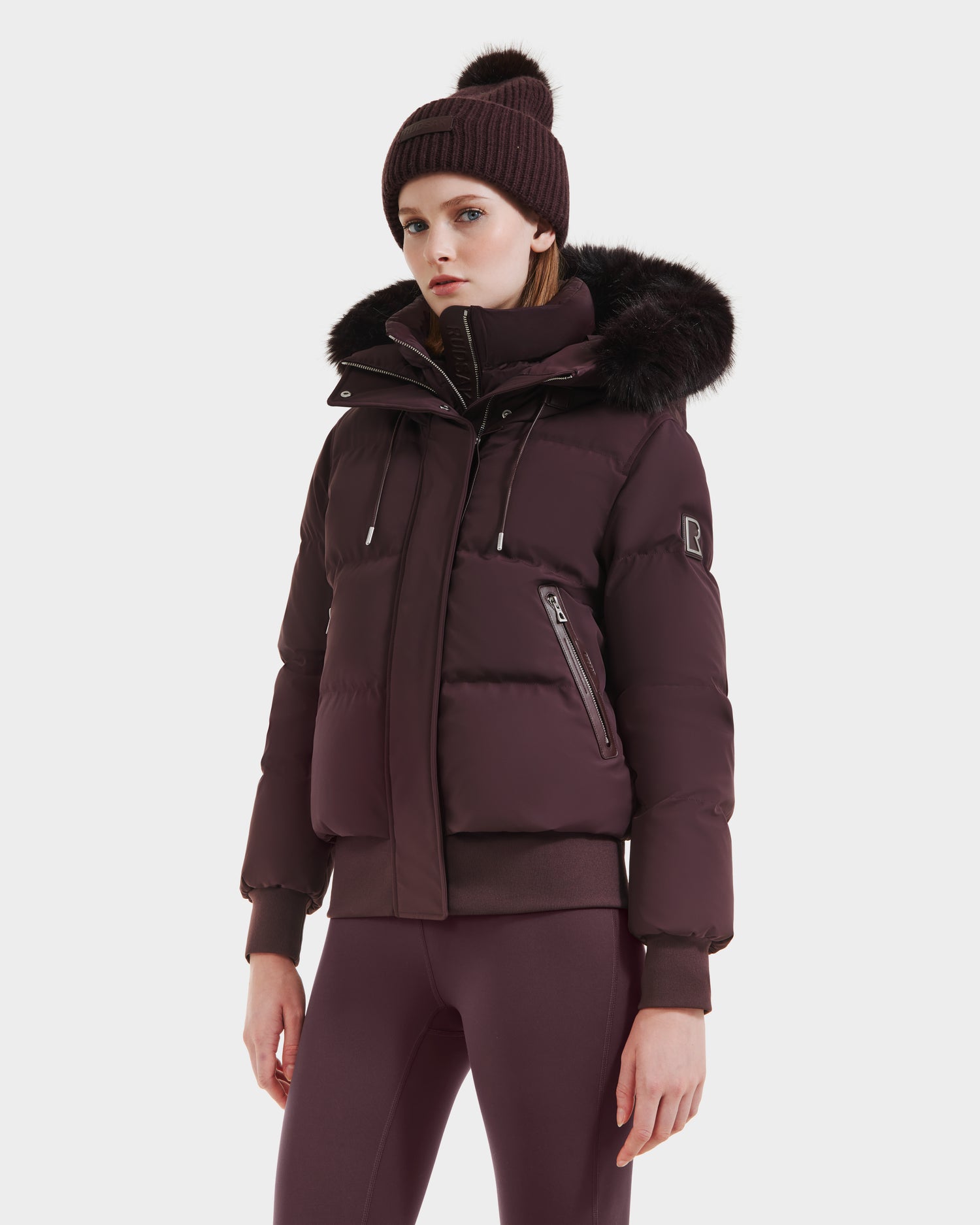 A woman in the Rudsak CIEYNA - 8125579 Bordeaux puffer jacket with recycled, weather-resistant shell and faux fur hood, paired with matching leggings and a knit pom-pom beanie, stands against a plain white background.