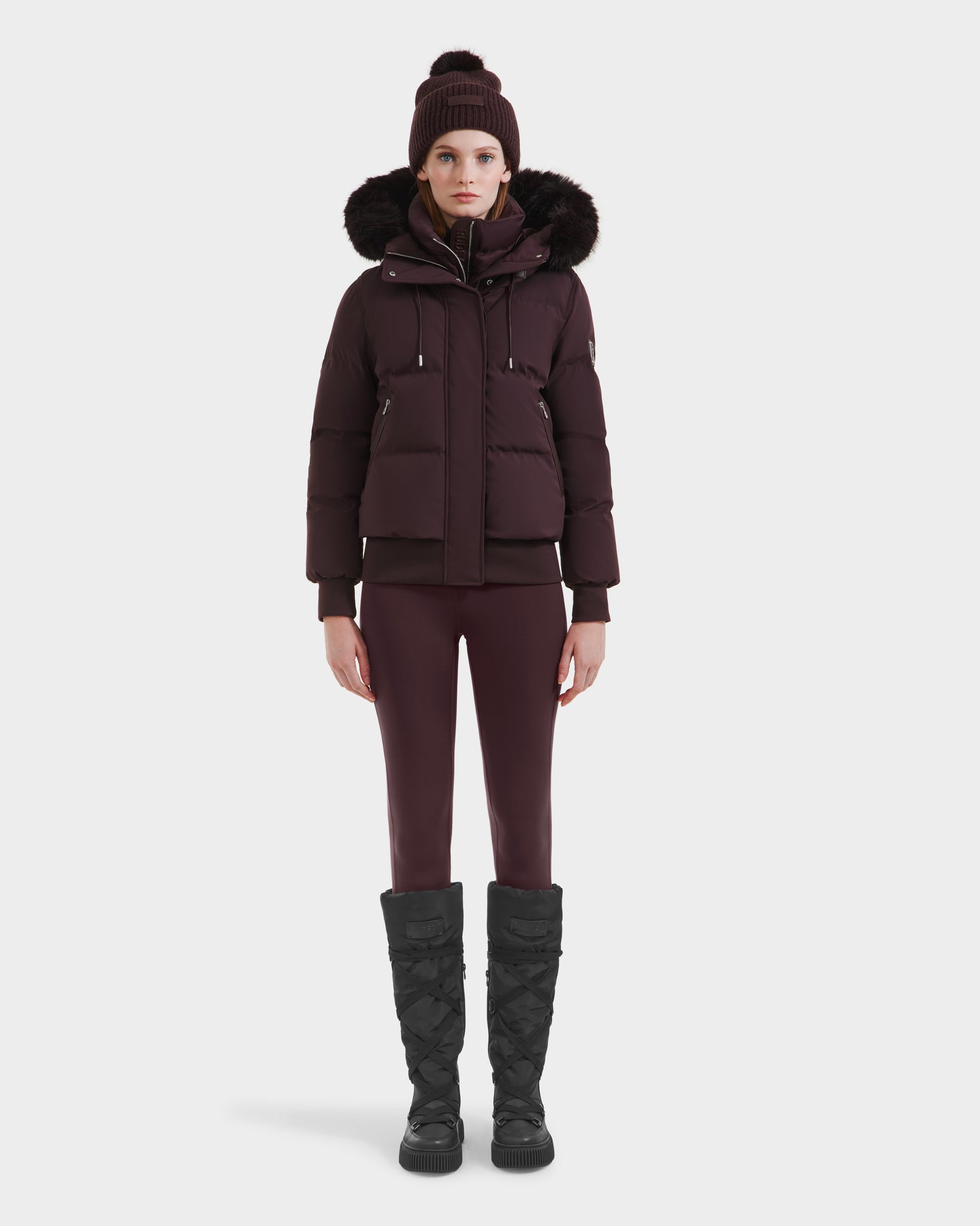 A person stands facing forward against a white background, wearing the Rudsak CIEYNA - 8125579 Bordeaux weather-resistant winter jacket with a fur-lined hood, matching beanie, fitted pants, and tall quilted black boots.