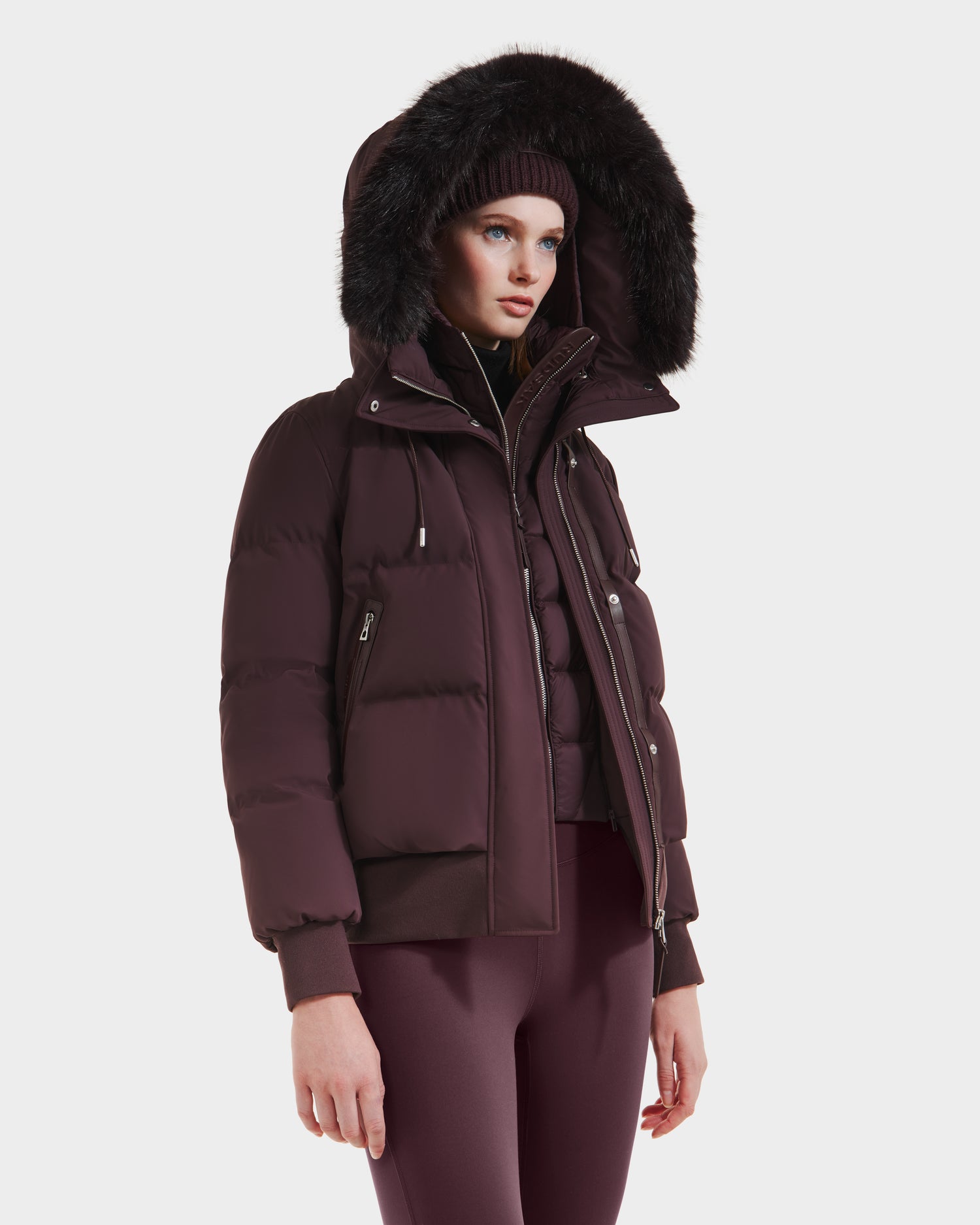 A woman wears the Rudsak CIEYNA - 8125579 Bordeaux weather-resistant puffer jacket with a fur-trimmed hood, matching leggings, and a knit beanie, standing against a plain white background.