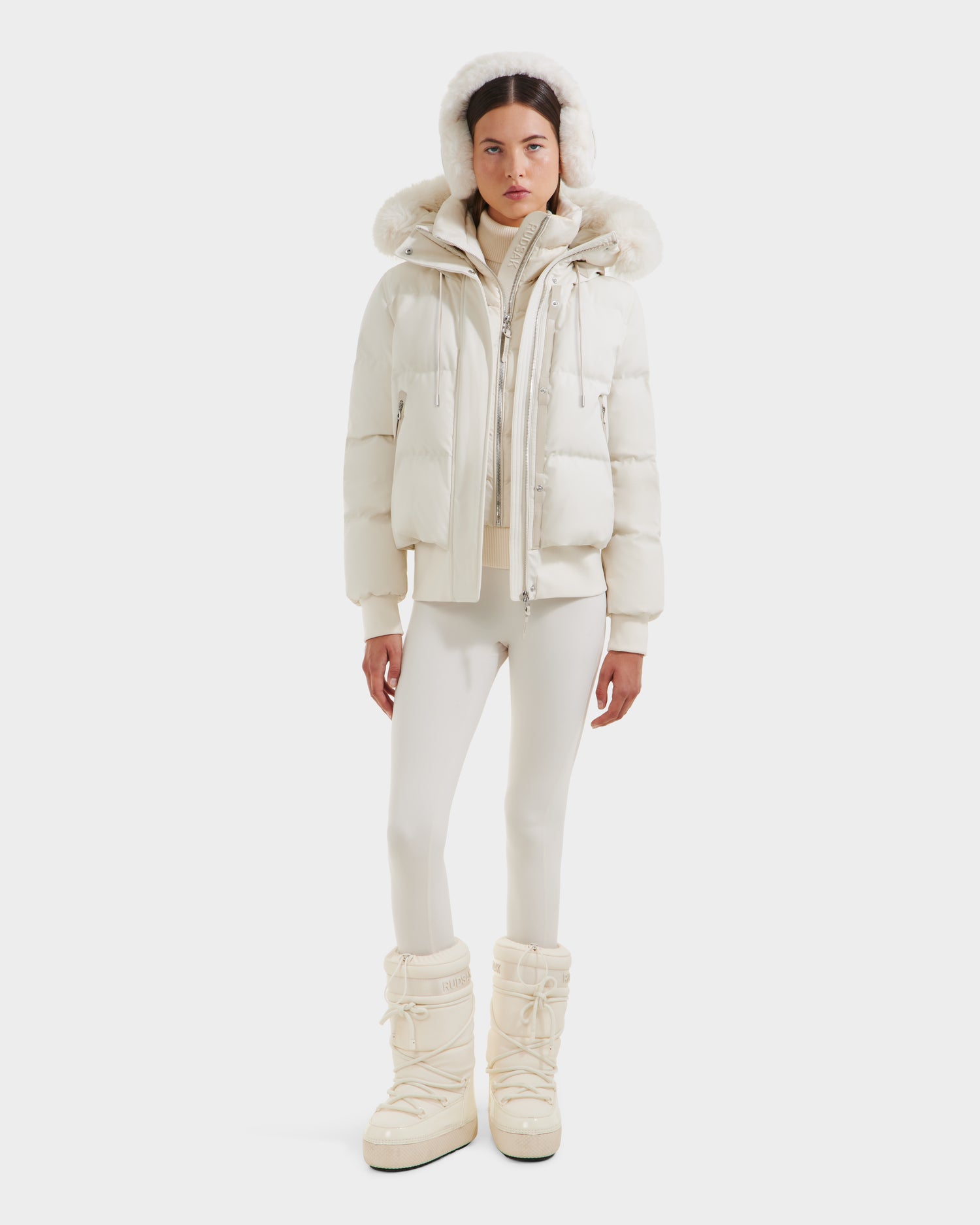 A woman stands against a white background wearing the Rudsak CIEYNA - 8125579 Vanilla down-filled bomber with a weather-resistant shell and faux fur hood, paired with white leggings and snow boots. Her expression is neutral, hands by her sides.