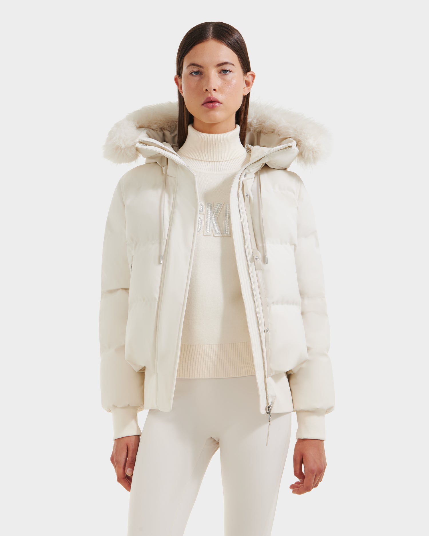 A woman in the Rudsak CIEYNA - 8125579 Vanilla white down-filled bomber with faux fur hood trim, a white turtleneck sweater, and white pants stands facing forward against a plain white background.