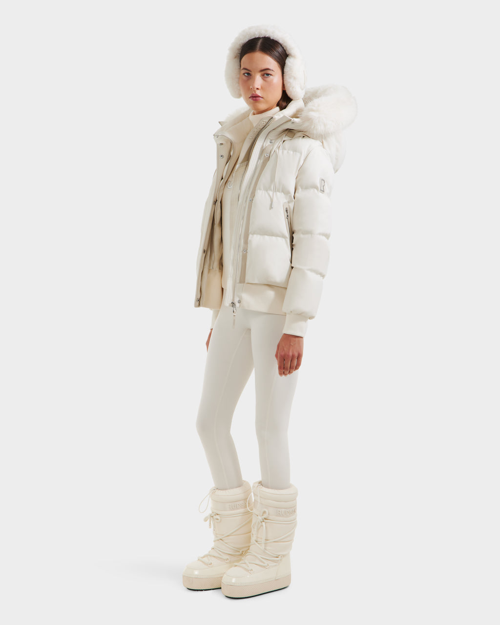 A person models the Rudsak CIEYNA - 8125579 Vanilla: a stylish all-white winter look with a weather-resistant puffy jacket, faux fur hood, fitted pants, and tall snow boots, set against a plain white background.