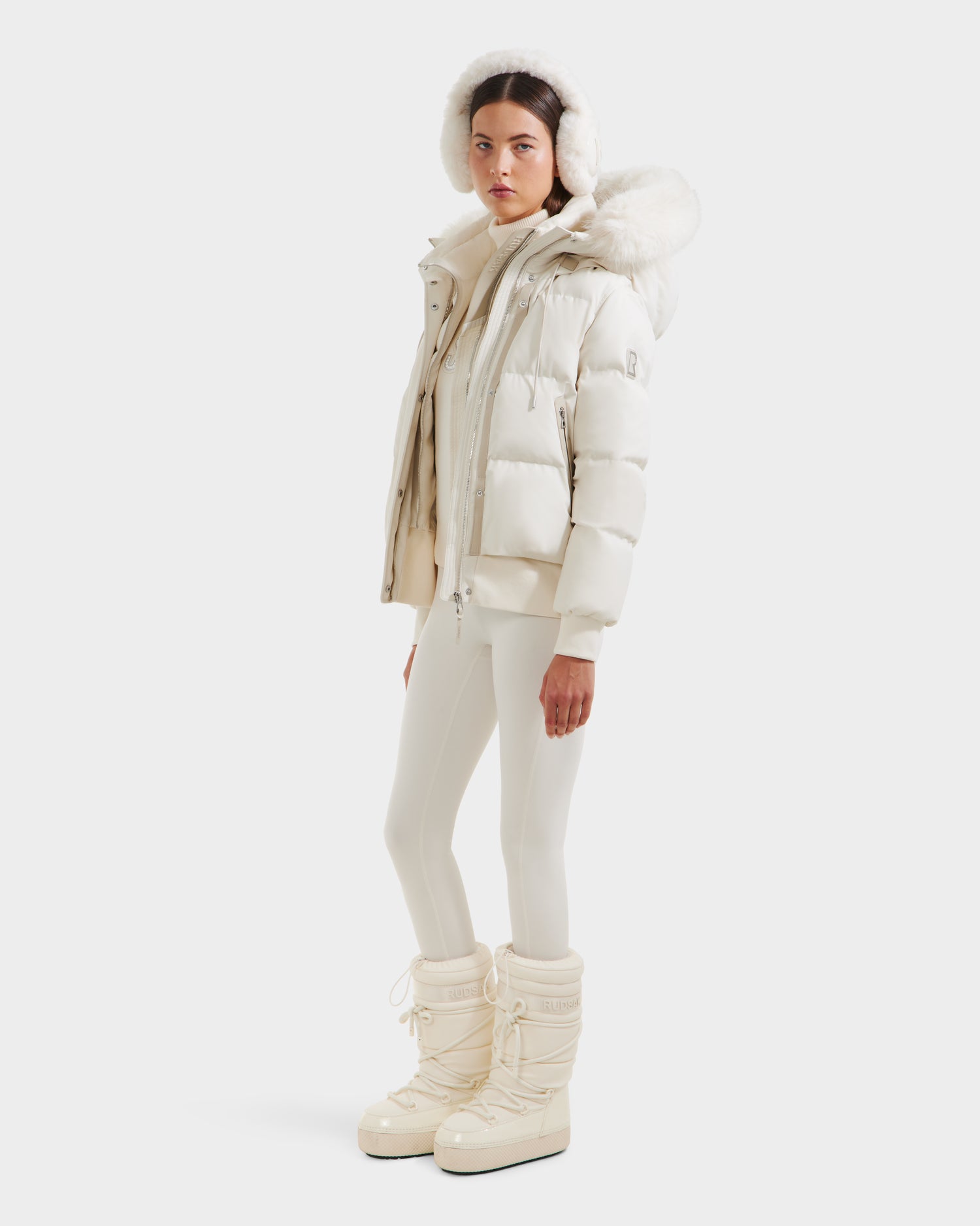 A person models the Rudsak CIEYNA - 8125579 Vanilla: a stylish all-white winter look with a weather-resistant puffy jacket, faux fur hood, fitted pants, and tall snow boots, set against a plain white background.