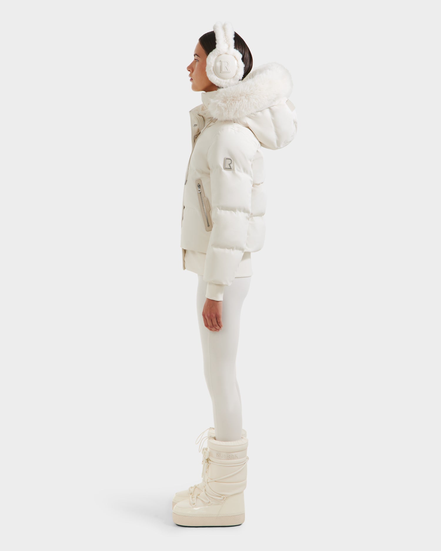 A woman in the Rudsak CIEYNA - 8125579 Vanilla down-filled bomber with a faux fur hood, white earmuffs, leggings, and snow boots stands in profile against a plain light background.