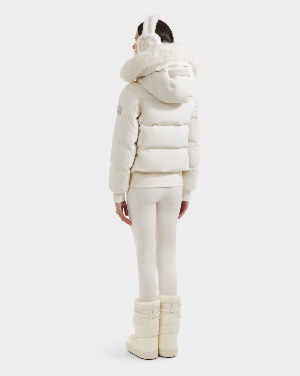 A person stands facing away in the Rudsak CIEYNA - 8125579 Vanilla down-filled bomber with faux fur hood, white leggings, earmuffs, and winter boots against a plain light background.