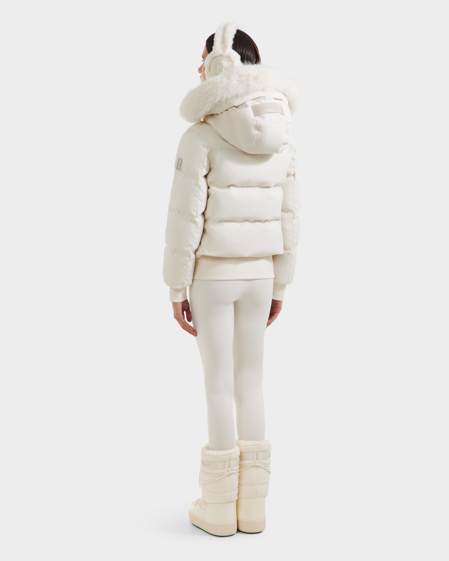 A person stands facing away in the Rudsak CIEYNA - 8125579 Vanilla down-filled bomber with faux fur hood, white leggings, earmuffs, and winter boots against a plain light background.