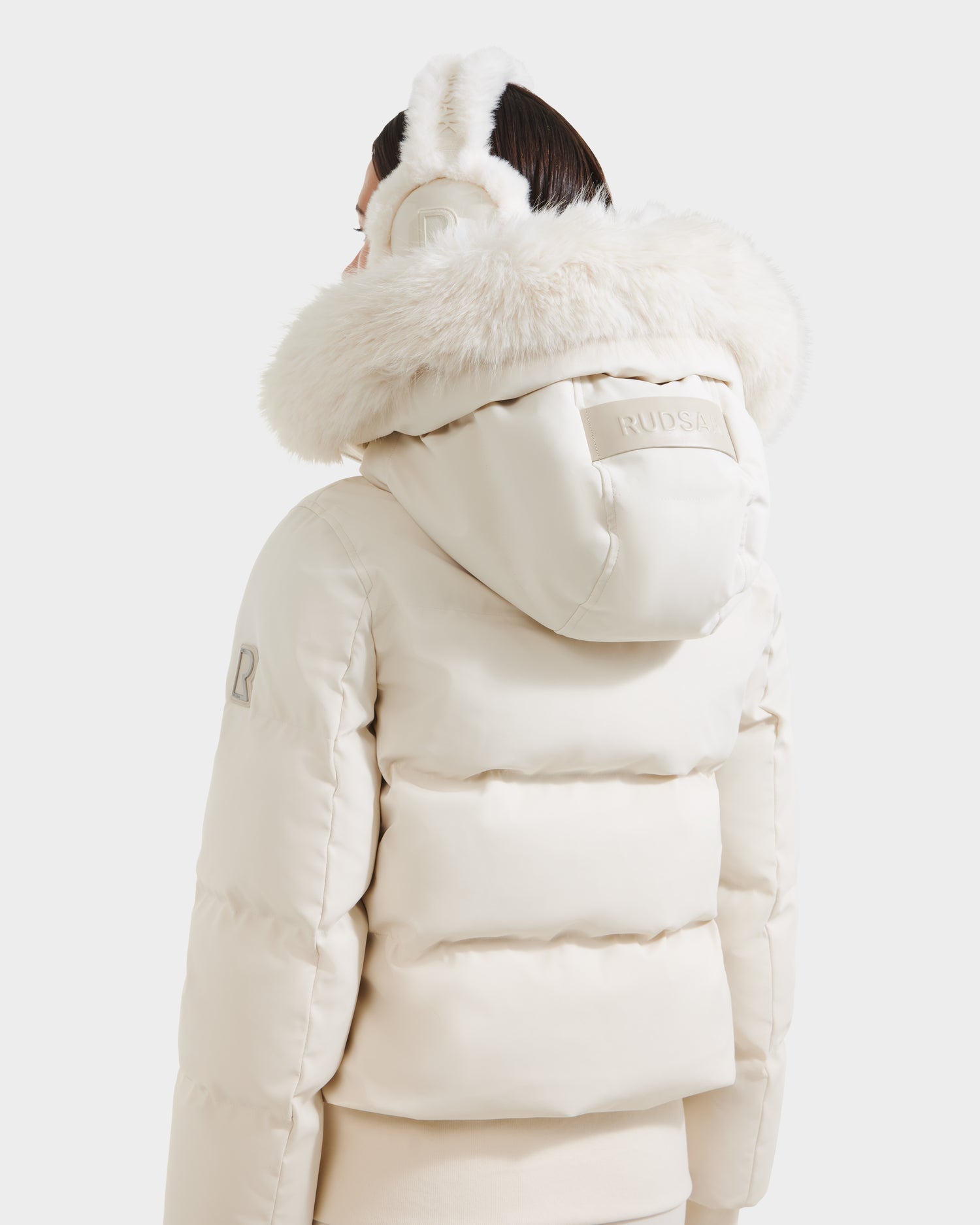 A person is seen from the back wearing the Rudsak CIEYNA - 8125579 Vanilla down-filled bomber jacket with faux fur hood and matching earmuffs, featuring a raised logo patch and weather-resistant shell, set against a white background.