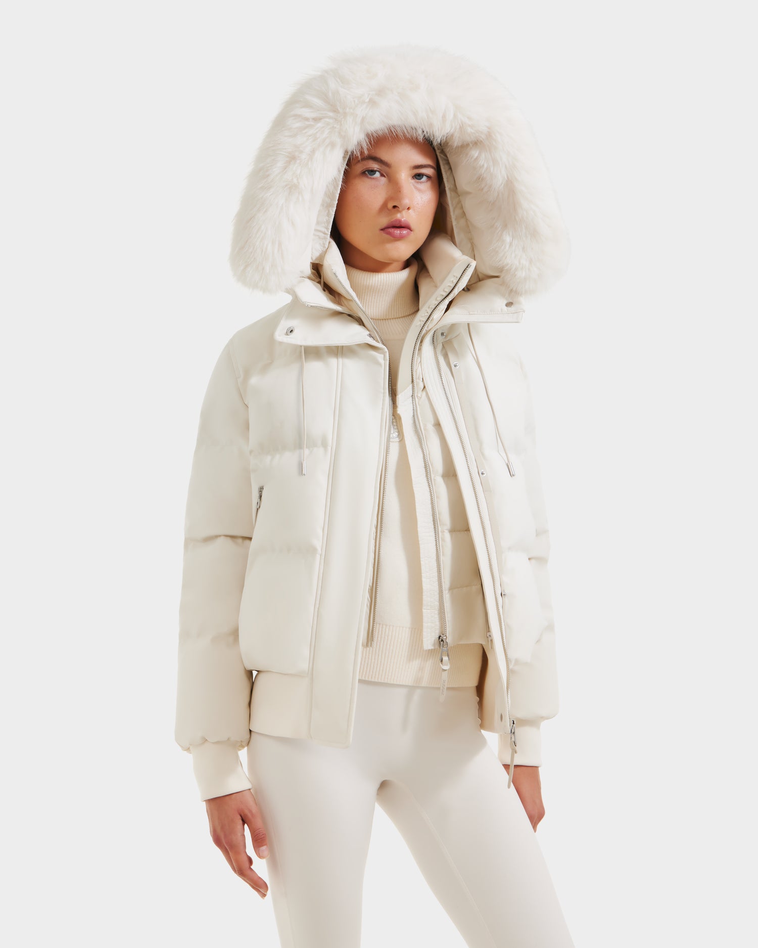 A person wearing the Rudsak CIEYNA - 8125579 Vanilla down-filled bomber with a large faux fur hood, beige turtleneck sweater, and white pants stands against a plain white background.