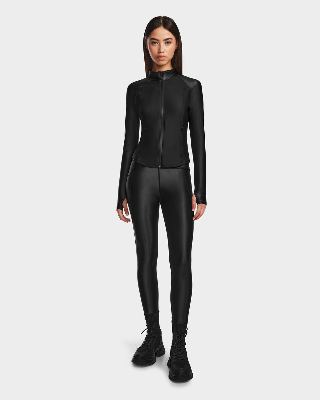 A woman faces forward against a white background, wearing Rudsaks CLARE - 8524519 Black fitted zip-up jacket and matching shiny stretch leggings with black lace-up boots.