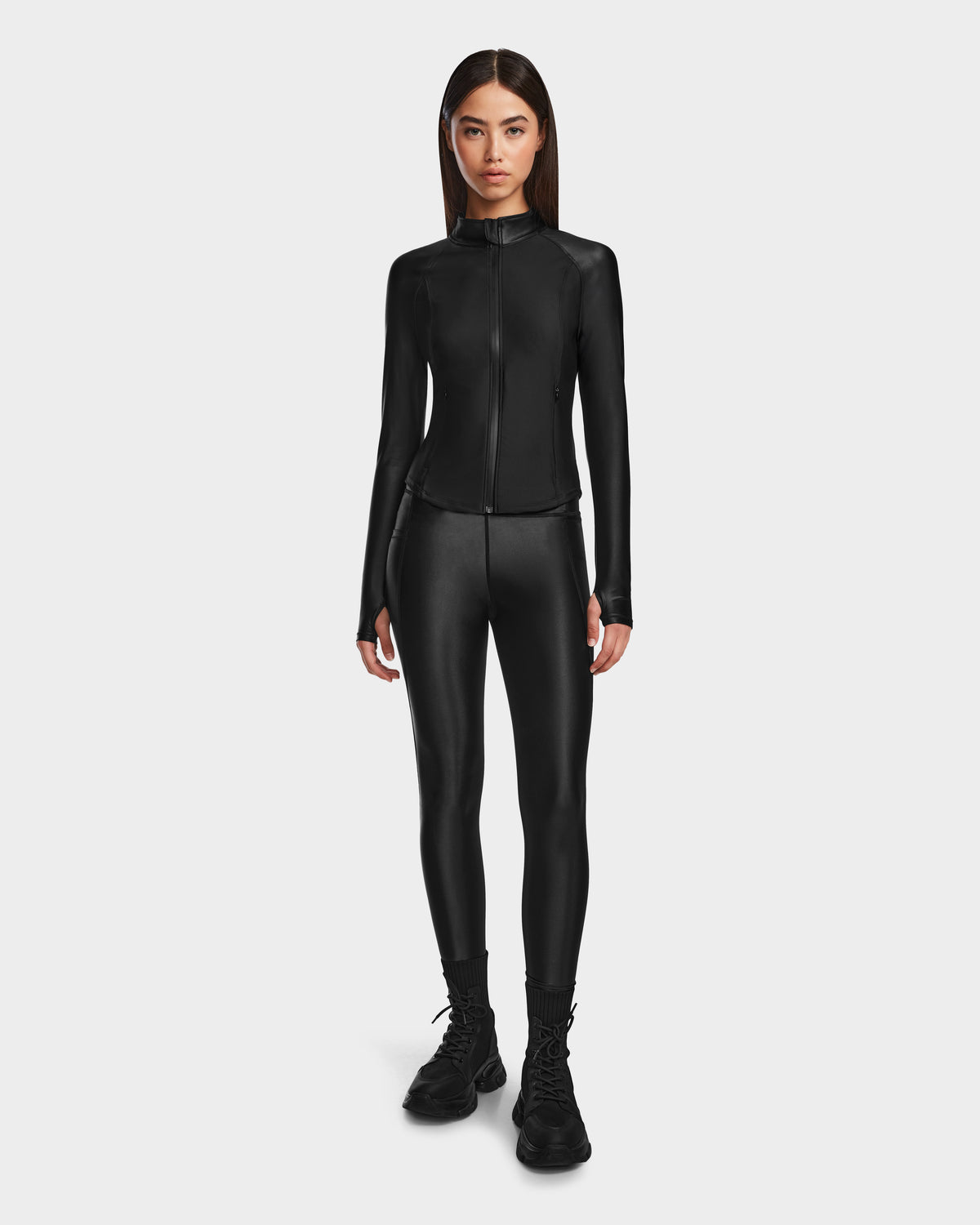 A woman faces forward against a white background, wearing Rudsaks CLARE - 8524519 Black fitted zip-up jacket and matching shiny stretch leggings with black lace-up boots.