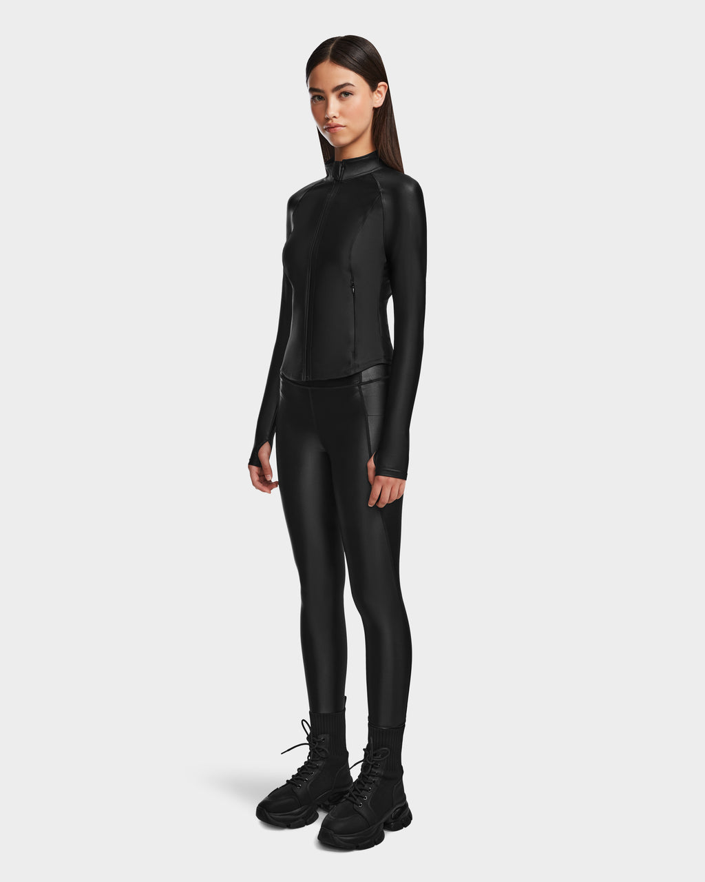 A woman faces forward in Rudsak’s CLARE - 8524519 Black fitted jacket and matching leggings with a shiny stretch finish, paired with black lace-up boots, set against a plain white background that highlights her silhouette.