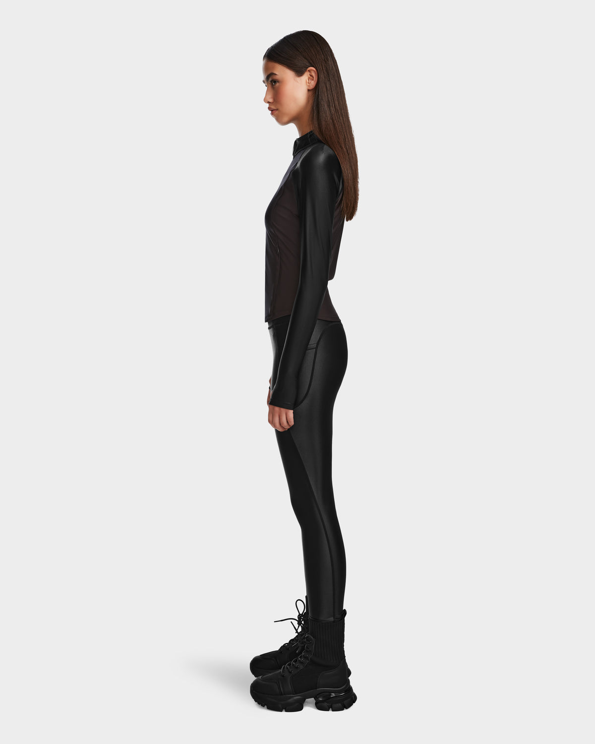A woman stands in profile against a plain background, wearing Rudsak’s CLARE - 8524519 Black long-sleeve top and matching leggings in shiny stretch fabric, paired with chunky black boots. Her straight brown hair is worn down.