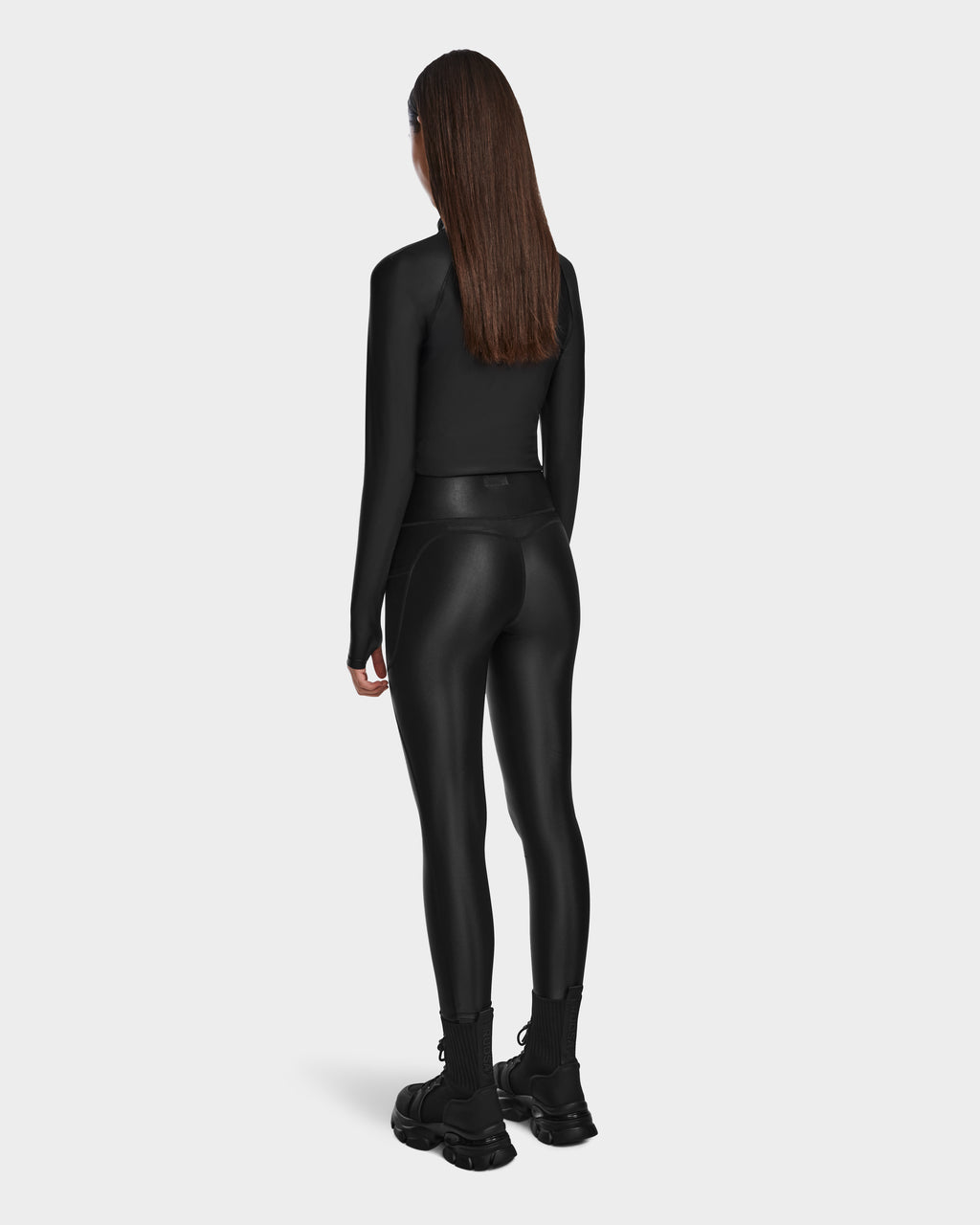 A woman with straight brown hair models the Rudsak CLARE - 8524519 Black long-sleeve top and shiny leggings, paired with chunky boots, her sculpted silhouette set against a plain light gray background.