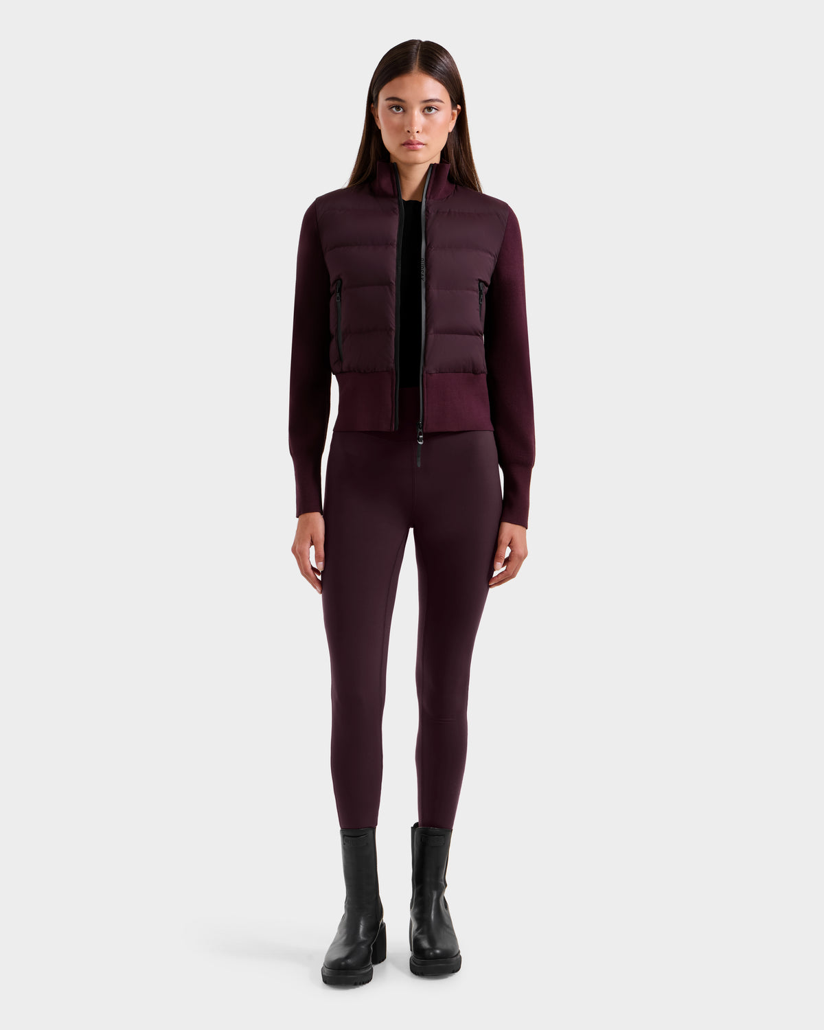 A woman faces forward against a plain light background, wearing the Rudsak CLAREMONT - 8125502 Bordeaux women’s hybrid down jacket for lightweight warmth, paired with matching leggings and black ankle boots.