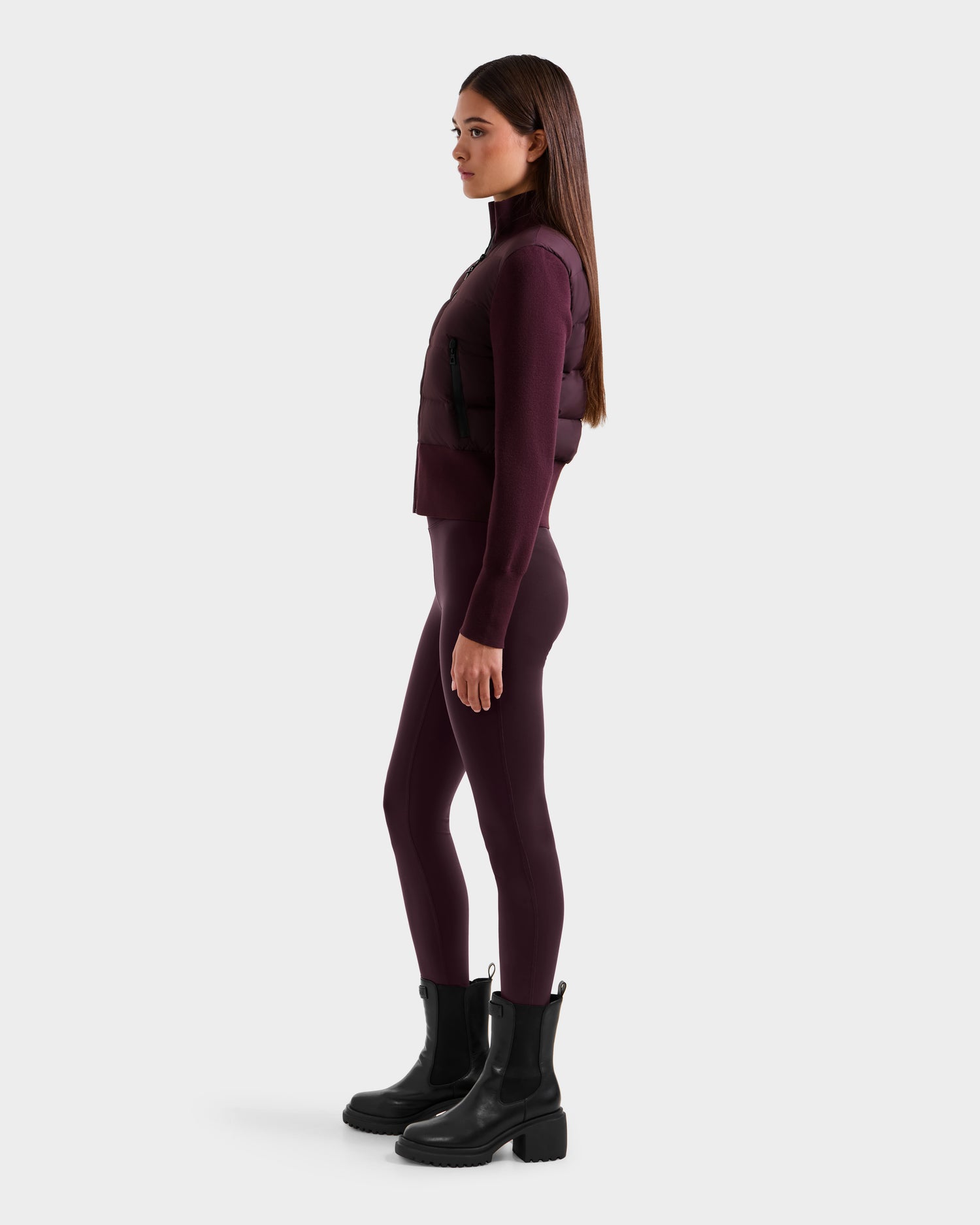 A woman stands in profile against a plain light background wearing the Rudsak CLAREMONT - 8125502 Bordeaux women’s short hybrid down jacket, paired with matching leggings, a long-sleeve top, and black ankle boots.