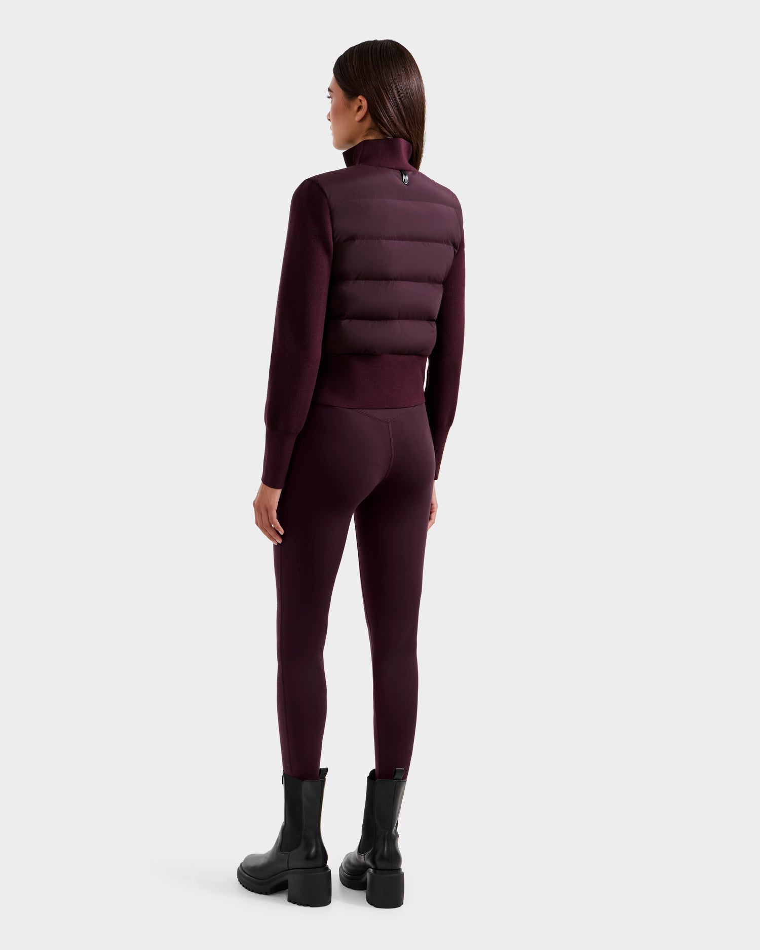 A woman with straight dark hair stands against a light gray background, wearing the Rudsak CLAREMONT - 8125502 Bordeaux hybrid down jacket, matching leggings, and black boots.