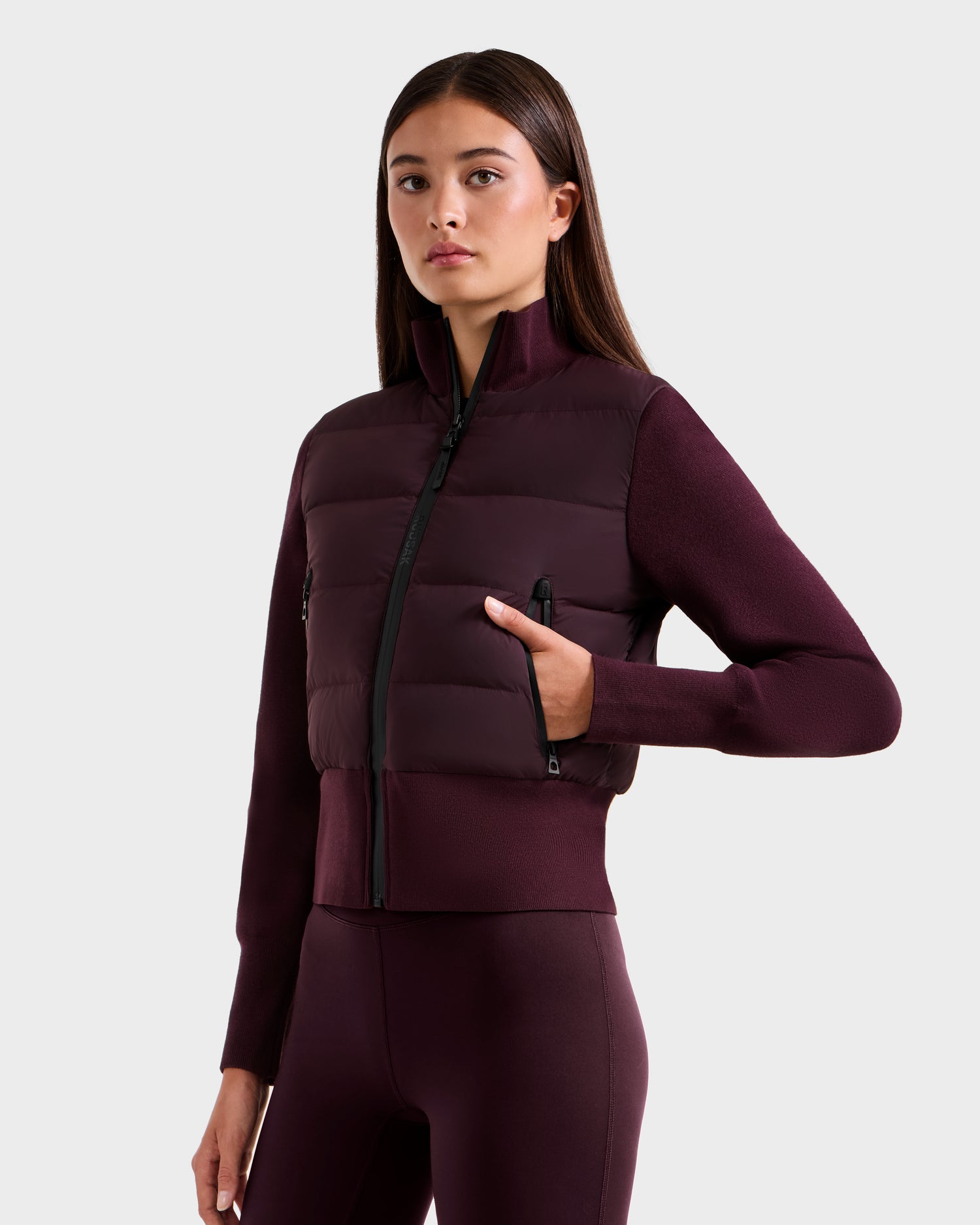 A woman with long brown hair faces forward, wearing the Rudsak CLAREMONT - 8125502 Bordeaux hybrid down jacket and matching pants, hand in pocket, against a plain light gray background, showcasing modern style and lightweight warmth.