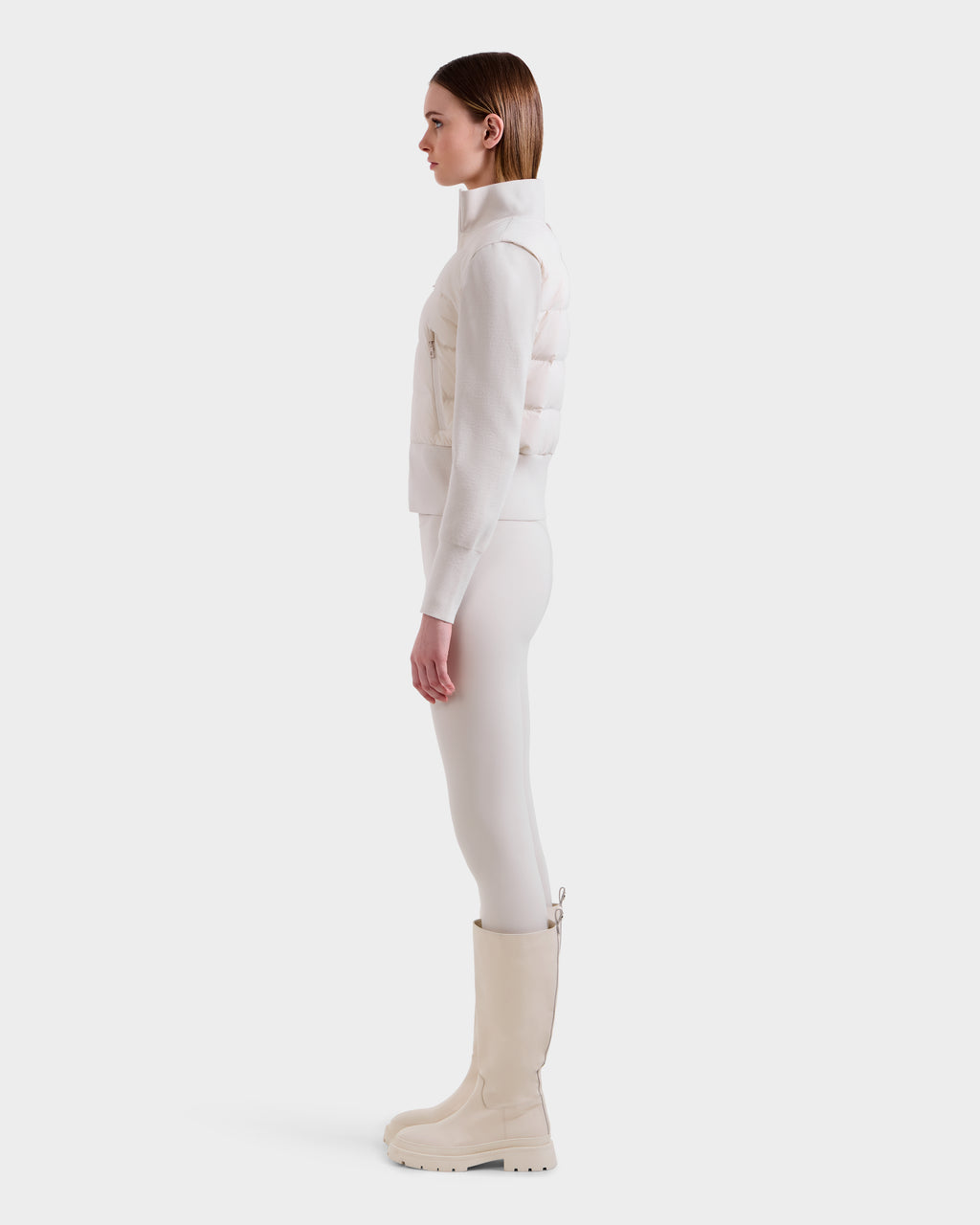 A woman stands in profile against a plain background wearing the Rudsak CLAREMONT - 8125502 Vanilla padded vest for lightweight warmth, paired with a light long-sleeve top, fitted pants, and tall off-white boots.