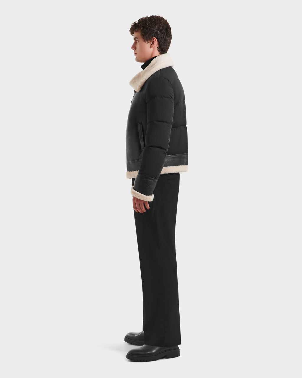 A man in profile wears the Rudsak CLAYTON - 6125501 Black down puffer jacket with white shearling trim, black pants, and black shoes, set against a plain white background.