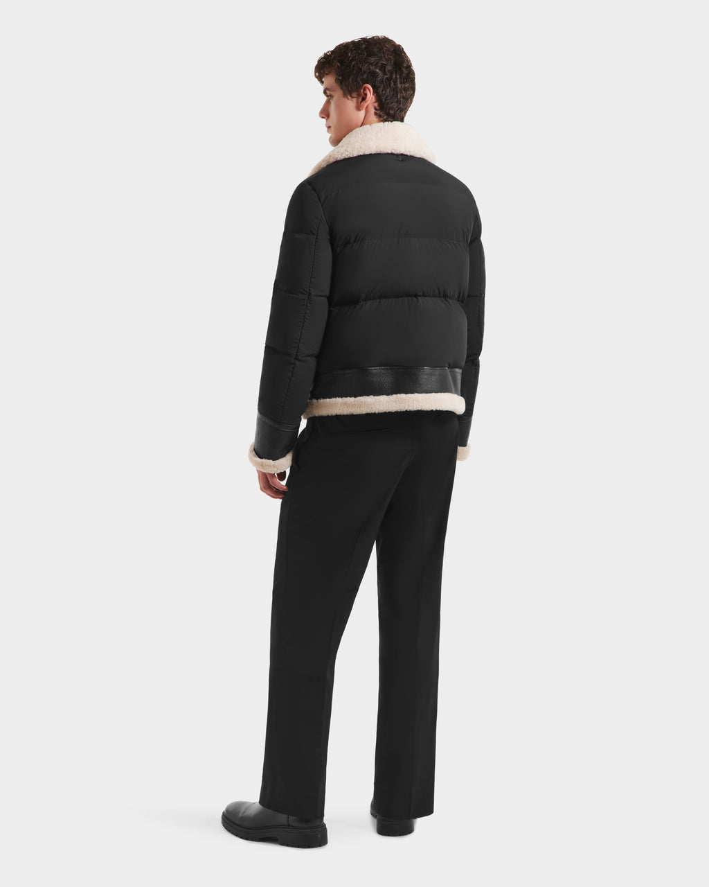 A person is seen from behind wearing the Rudsak CLAYTON - 6125501 Black down puffer jacket with a light shearling collar and trim, paired with black pants and shoes, standing against a plain white background.