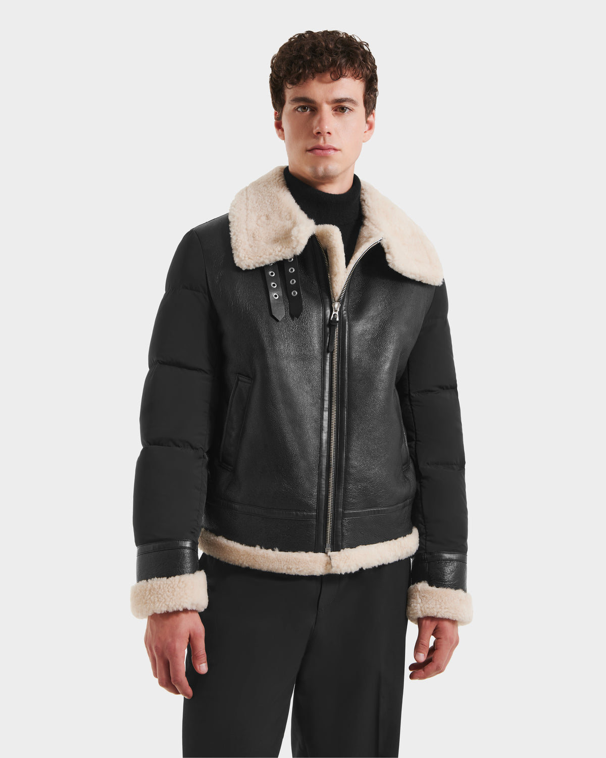 A man in the Rudsak CLAYTON - 6125501 Black men’s shearling jacket with a wide beige fur collar and cuffs, black trousers, and a black turtleneck stands against a plain white background.