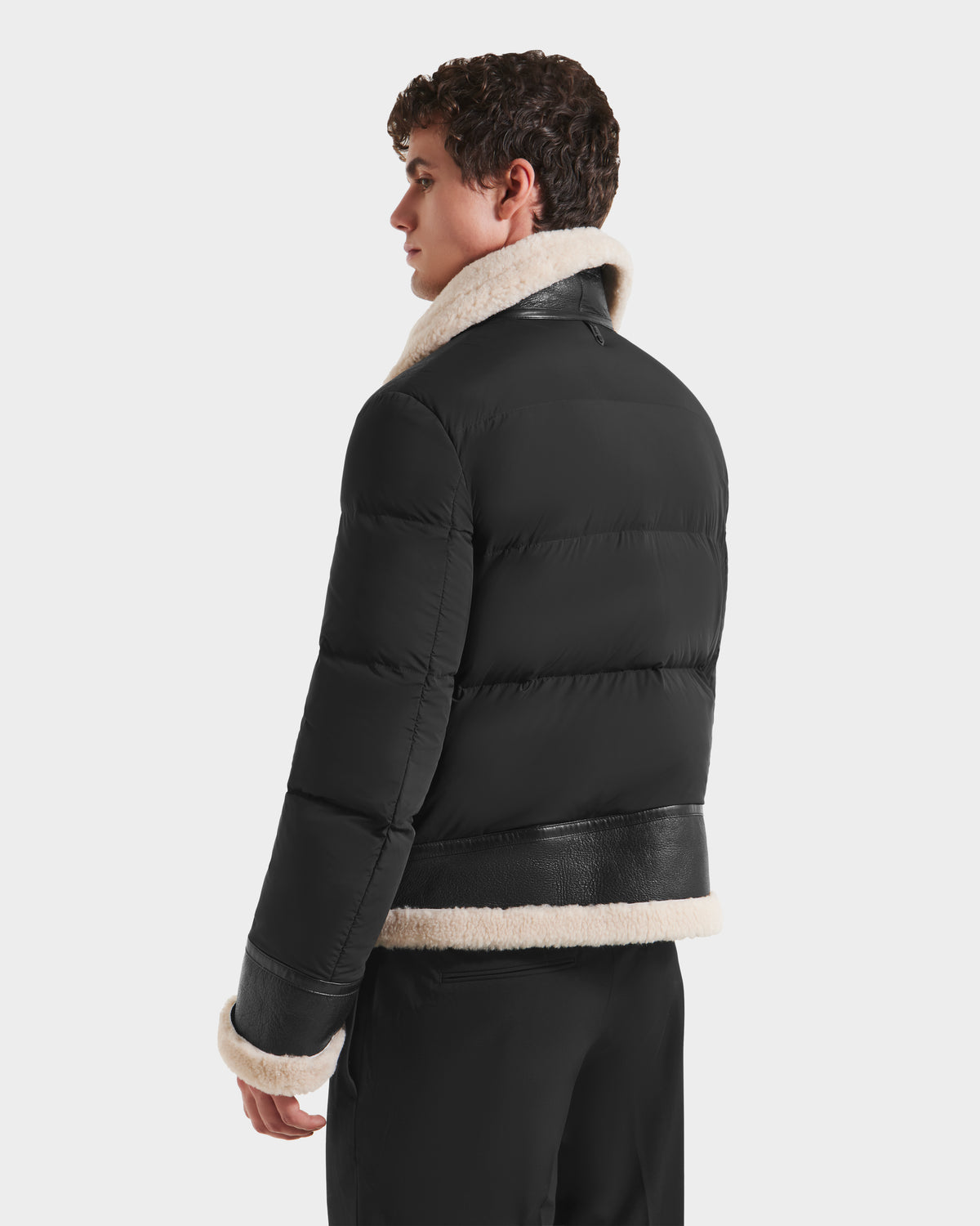 A young man with short curly hair models the Rudsak CLAYTON - 6125501 Black men’s down puffer jacket, featuring a light beige shearling collar and cuffs, as he looks over his shoulder against a plain light background.