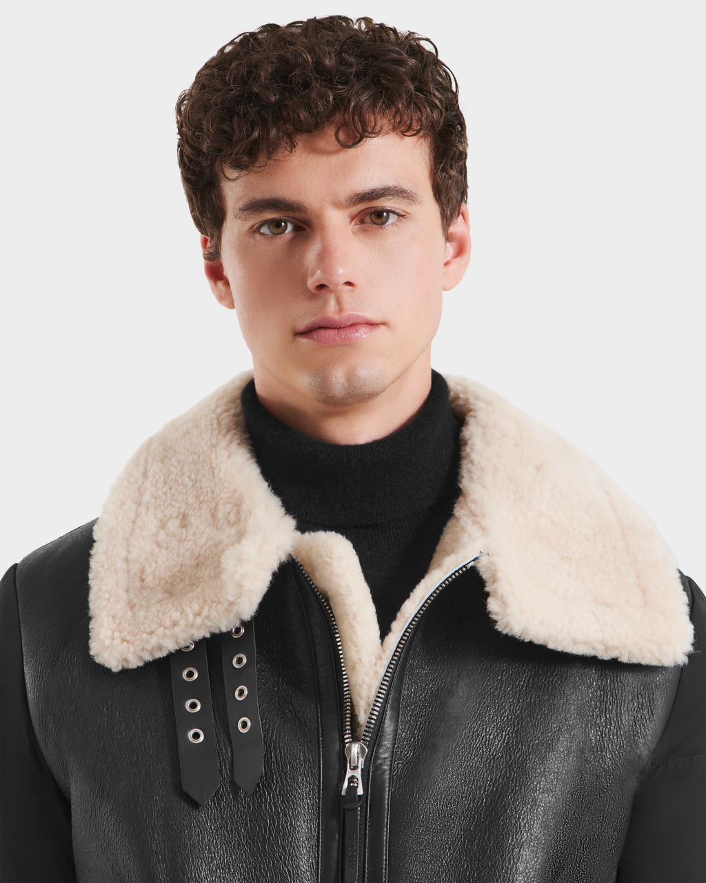 A young man with short curly brown hair wears the CLAYTON - 6125501 Black shearling jacket by Rudsak, featuring a large beige collar over a black turtleneck, standing against a light background.