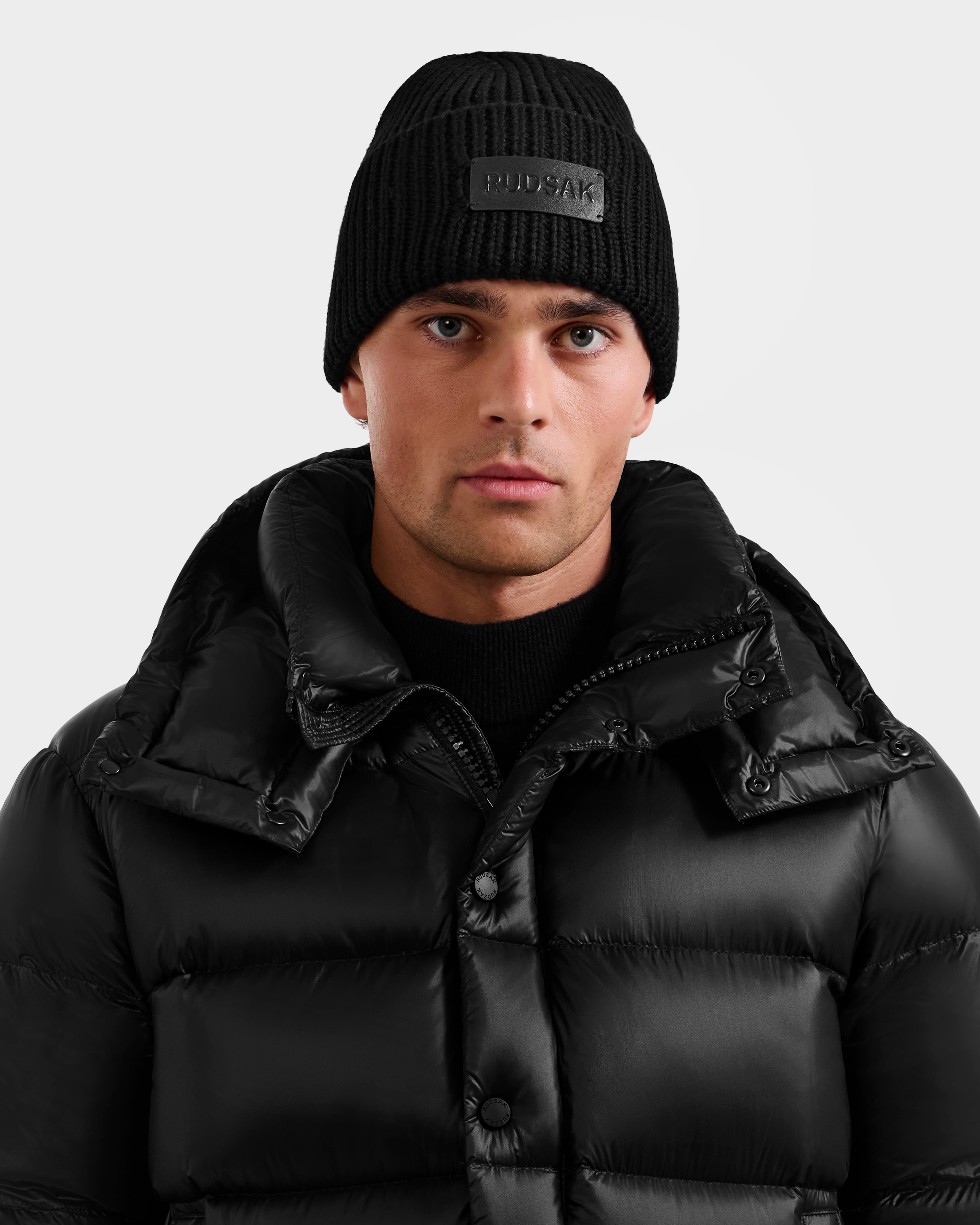 A man wearing a black Rudsak COLIN - 9424518 puffer jacket and a matching beanie looks straight at the camera against a plain light background.