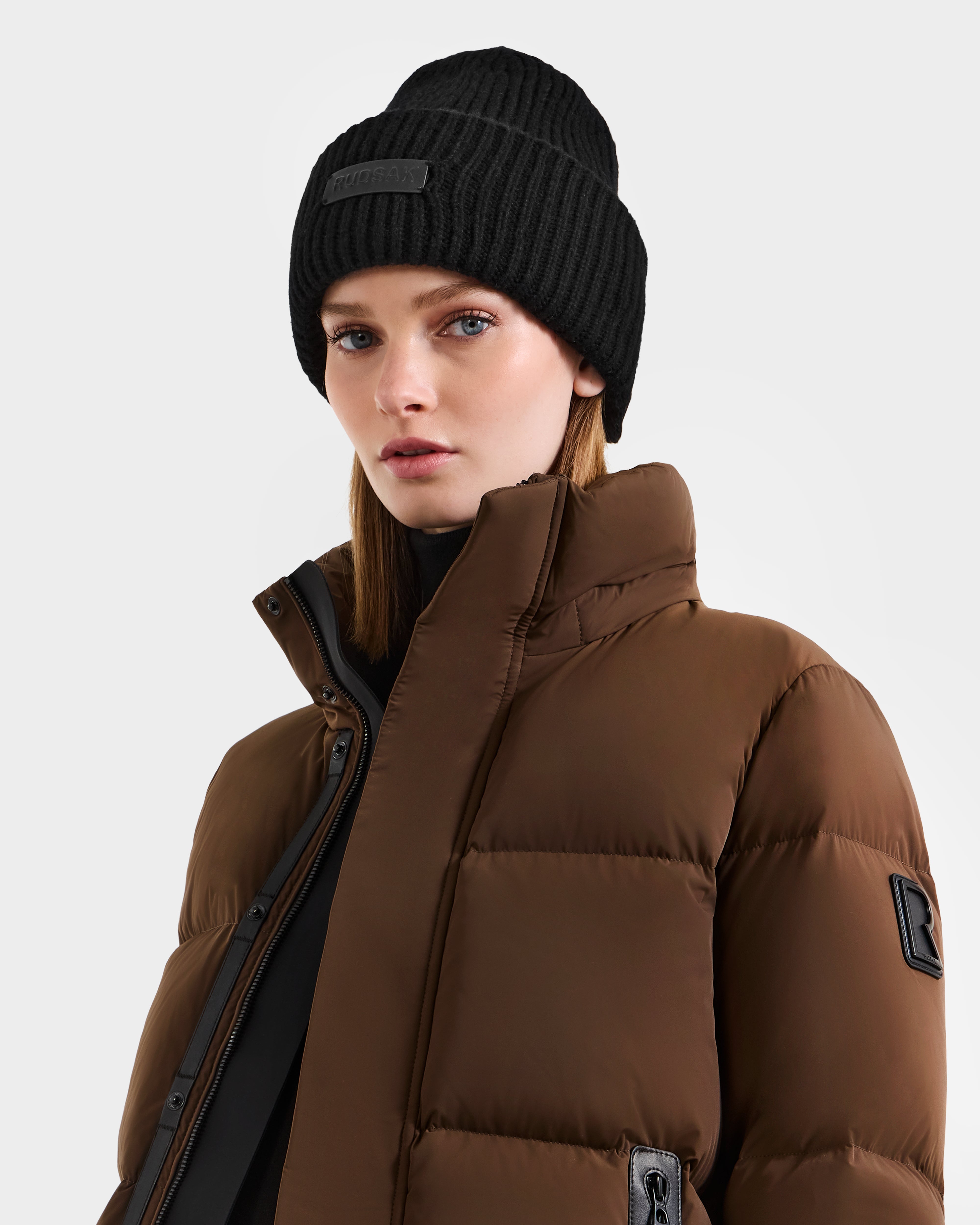 A person wears the Rudsak COLIN - 9424518 Black hat and a brown puffer jacket with a high collar, gazing confidently at the camera against a plain light background.