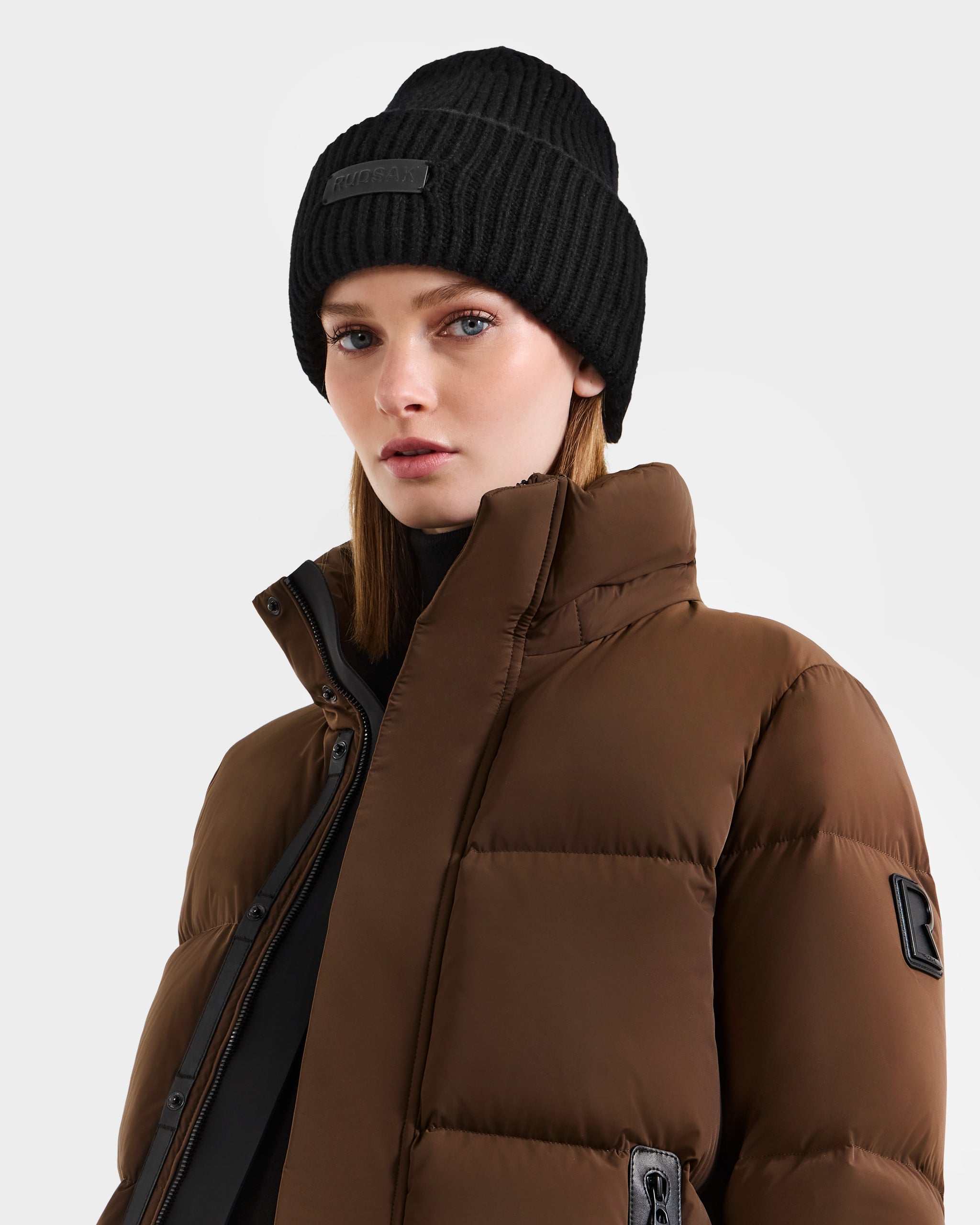 A person wears the Rudsak COLIN - 9424518 Black hat and a brown puffer jacket with a high collar, gazing confidently at the camera against a plain light background.