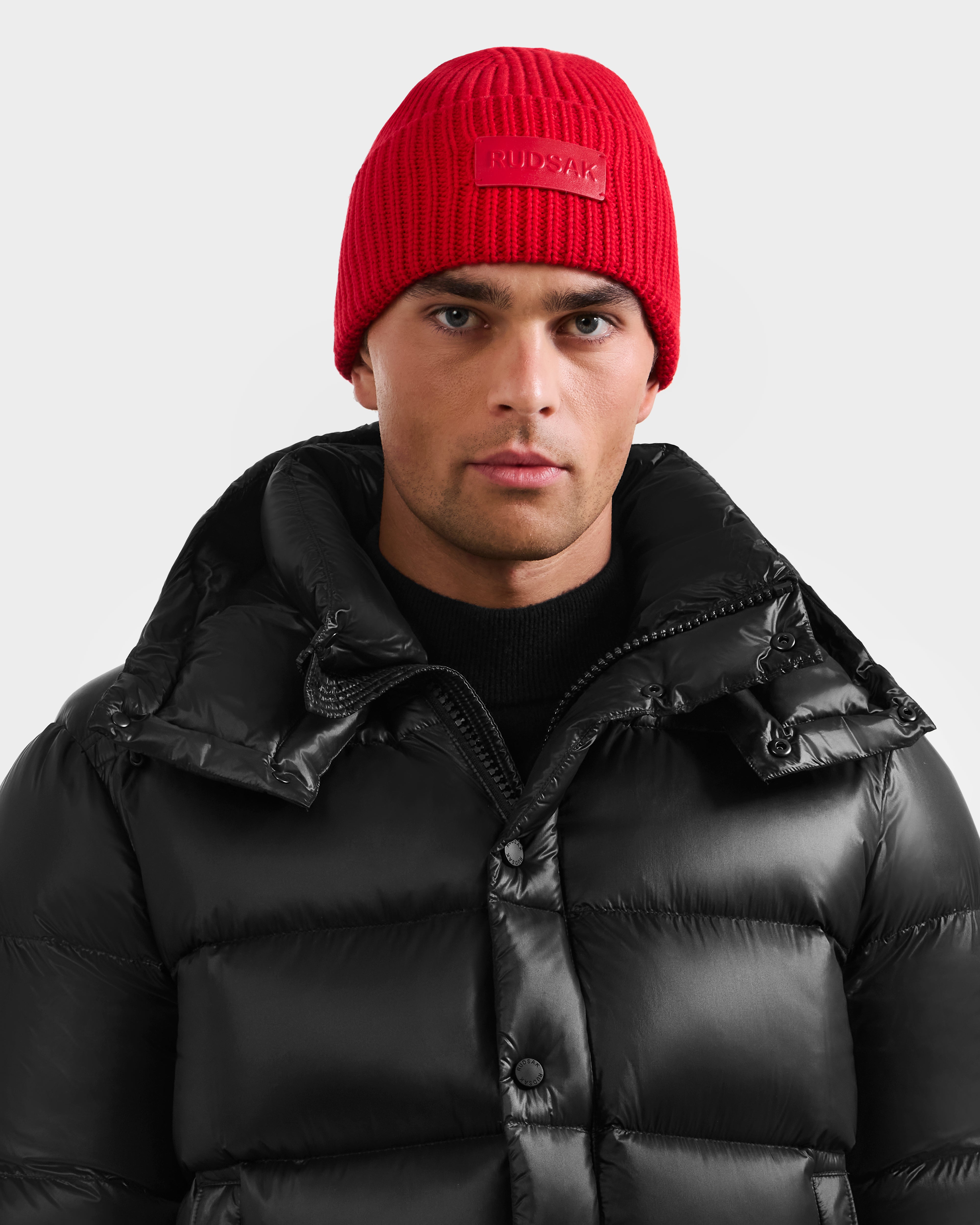 A man wears the Rudsak COLIN - 9424518 Flame Red beanie and a black high-collar puffer jacket, looking directly at the camera against a plain light background.