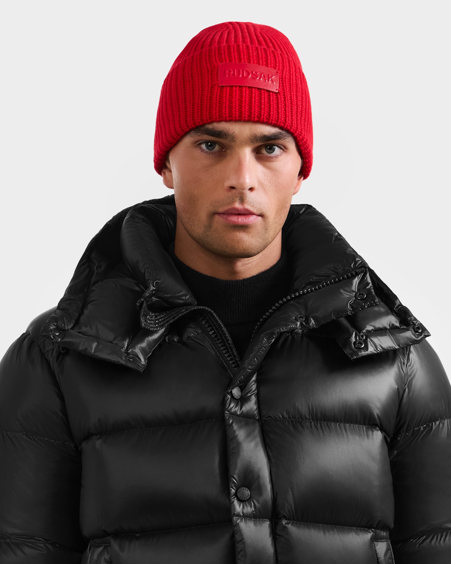 A man wears the Rudsak COLIN - 9424518 Flame Red beanie and a black high-collar puffer jacket, looking directly at the camera against a plain light background.